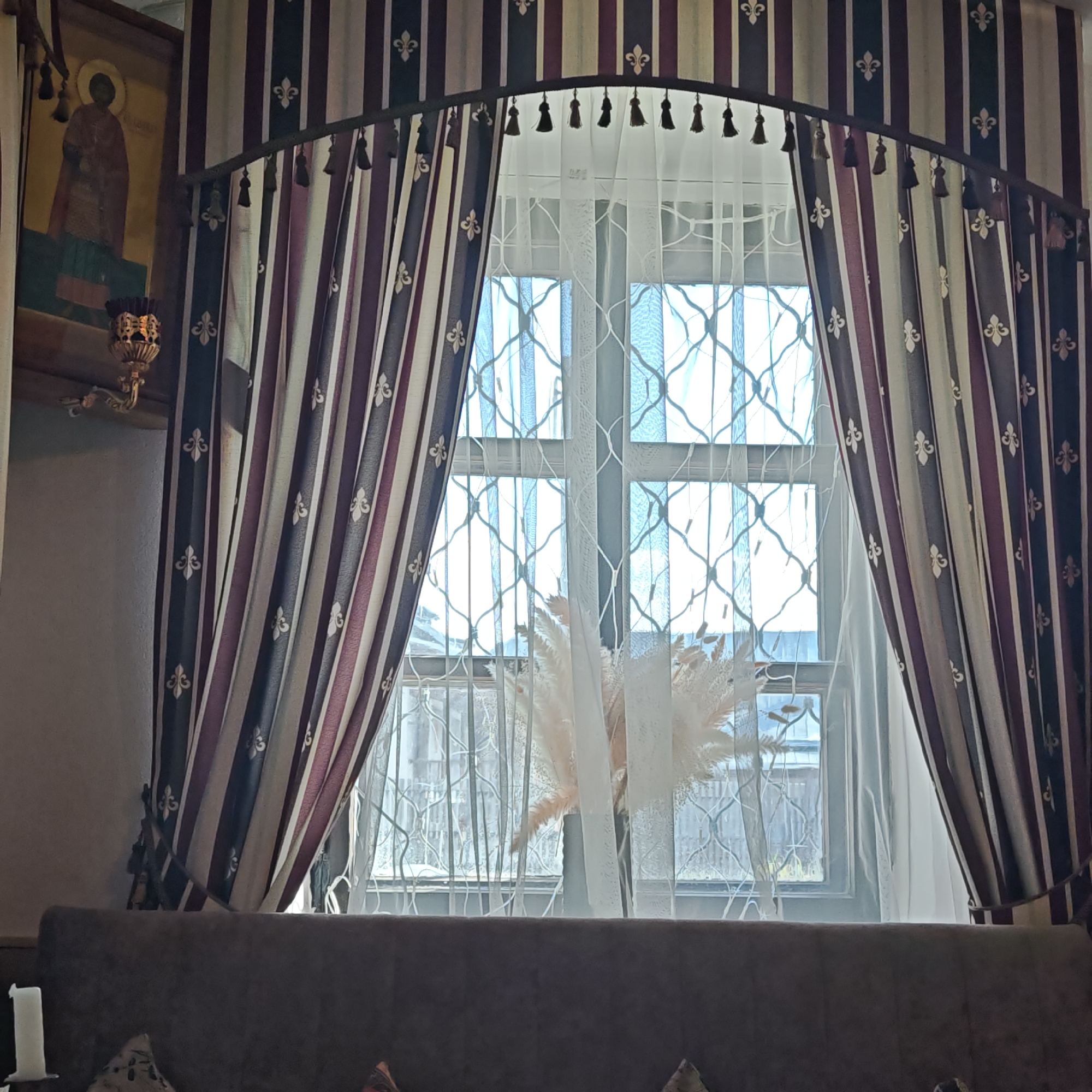 The image depicts an interior view, focusing on a window adorned with striped curtains featuring a fleur-de-lis pattern and tassel trim. The window itself has a decorative lattice design. Sheer white curtains filter the light, and a vase containing feathery plants sits on the windowsill. To the left of the window, a religious icon is visible, accompanied by an ornate sconce. The bottom of the frame includes the top of a sofa or similar seating. Based on context clues, the location is likely Rostov, Russia.