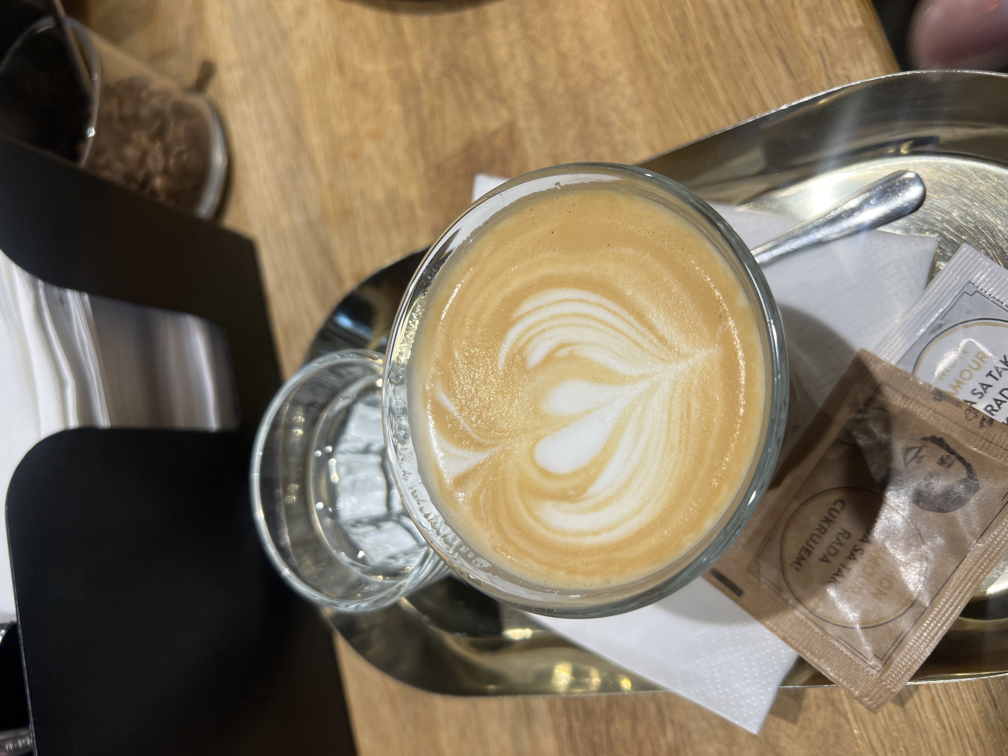 A close-up, eye-level shot shows a latte in a clear glass mug on a metal tray, positioned on a wooden table. The latte has a heart design in the foam. To the left of the latte is a glass of water. In front of the latte is a white napkin and a brown sugar packet with text in an unknown language. A silver spoon rests on the tray to the right of the latte. In the background, a black napkin dispenser holds white napkins, and a glass container filled with coffee beans is visible. The overall setting appears to be a cafe in Vysoké Tatry, Slovakia.