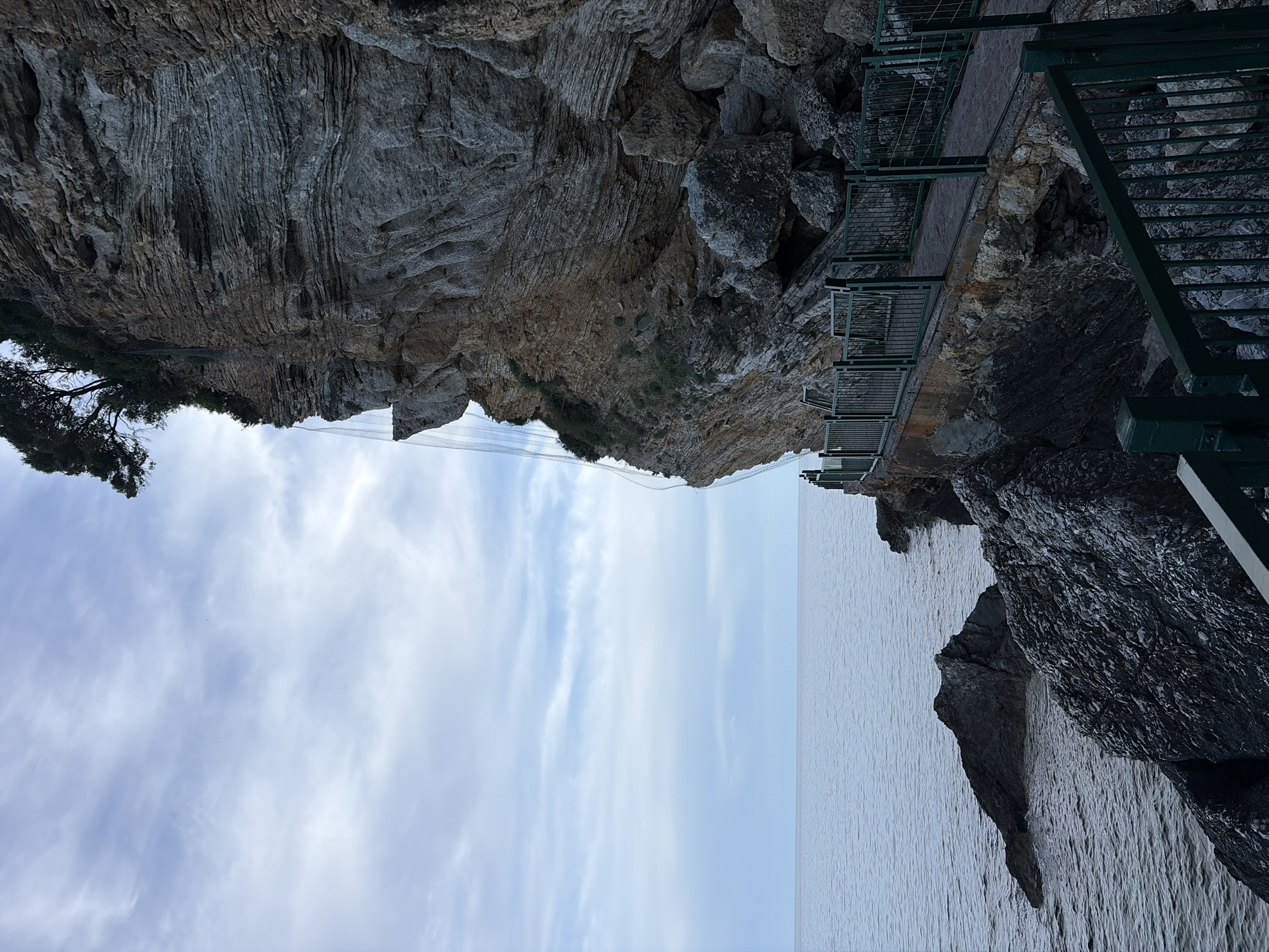 The image depicts a coastal scene in Budva, Montenegro, oriented with the land appearing above and the sky and sea below. A weathered, stratified cliff face occupies the upper portion of the image, transitioning from gray rock at the top to a tan and rocky section lower down. A wire mesh safety barrier is visible along the cliff edge. A metal stairway and walkway cling to the side of the cliff.
The lower portion of the image shows a pale blue sky with wispy clouds and calm, light gray sea. A dark gray rock formation rises from the sea on the right side of the image, adjacent to the stairway. The scene suggests a constructed path along a rugged coastline.