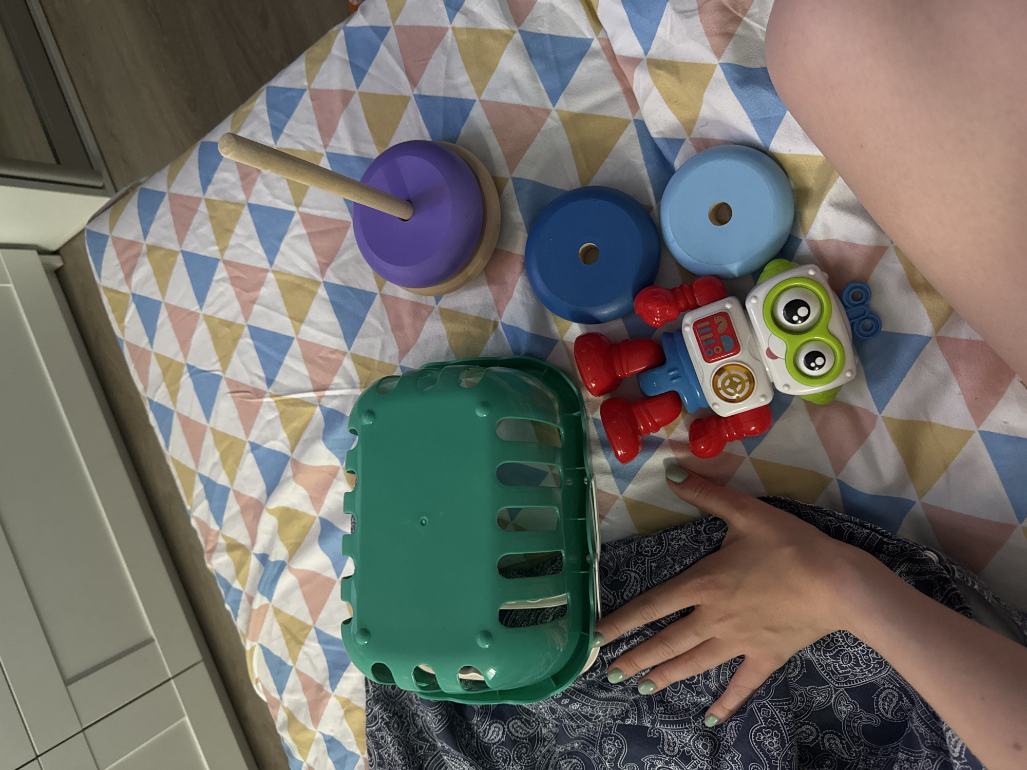 A top-down shot shows a collection of toys on a patterned surface, possibly a bedspread, next to a person’s leg and arm. The toys include a wooden stacking toy with purple and natural wood pieces, blue circular stacking rings, a robot toy, and a green plastic basket. The person’s hand rests near the basket and the robot toy. The setting appears to be an interior space, with a portion of a cabinet or wall visible on the left side of the image. It is not possible to determine the location as Moscow, Russia from the given visual information.