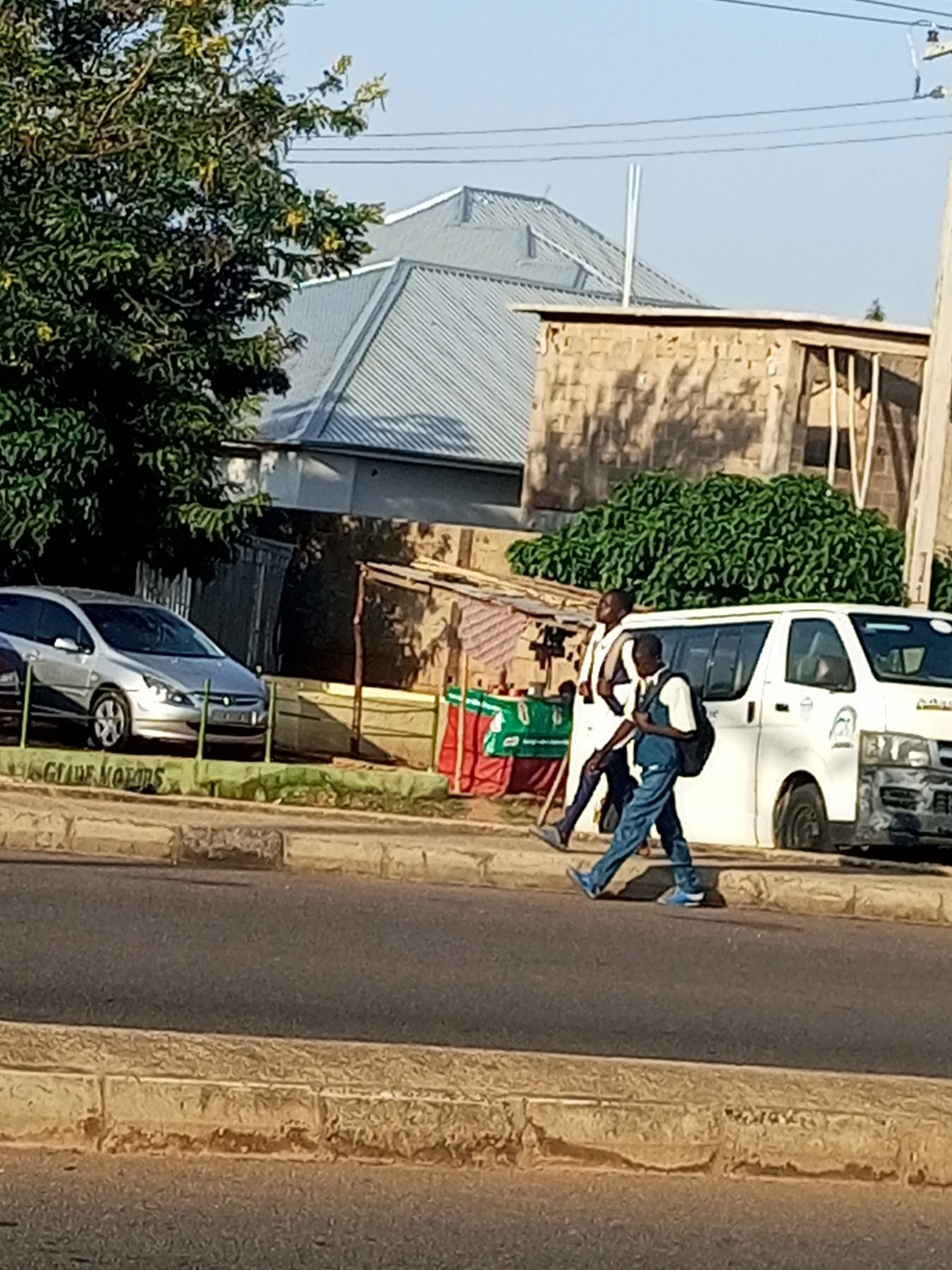 In Garko, Nigeria, two young males are captured mid-stride on a paved road. The individual on the left, wearing a white shirt and dark trousers, carries a bag over his left shoulder. The male on the right is dressed in a blue shirt and blue jeans, also with a bag on his shoulder. They are walking past a white van positioned on the side of the road. Adjacent to the van is a low structure with red and green coverings. In the background, a silver car is parked behind a green barrier. A mature tree with green foliage and yellow accents stands to the left of the scene, with buildings and a corrugated metal roof visible behind it. The environment appears to be outdoors with clear skies overhead.