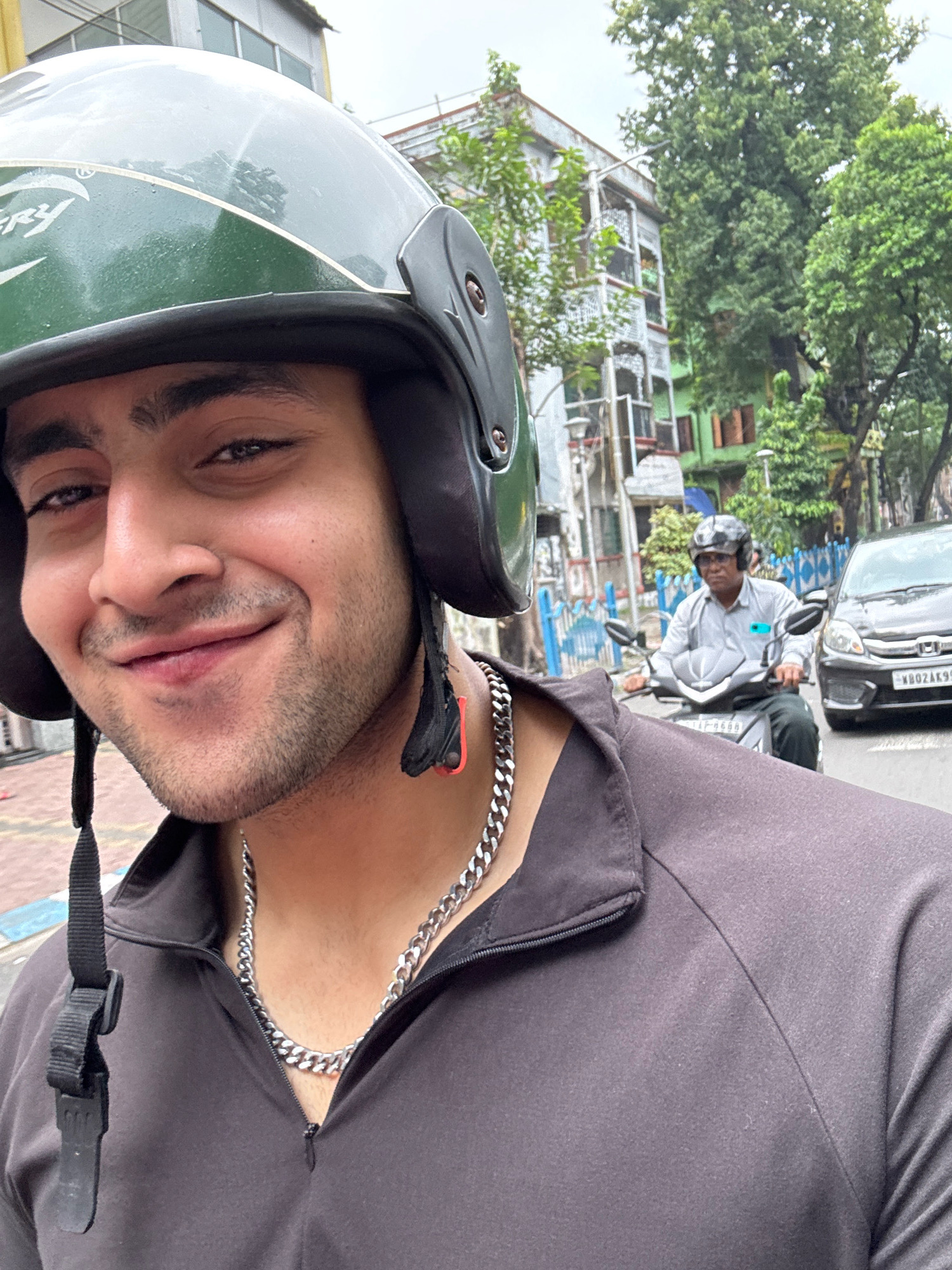 A man wearing a green helmet and a dark gray zip-up jacket smiles at the camera, taking a selfie from a motorcycle. He has dark hair, brown eyes, and a stubble beard. A thick silver chain is visible around his neck. In the background, another man on a motorcycle wears a light gray shirt and dark pants, with headphones on. A black car is parked on the right side of the road. The scene appears to be a street in Kolkata, India, with buildings and lush green trees lining the road. The sky is overcast.