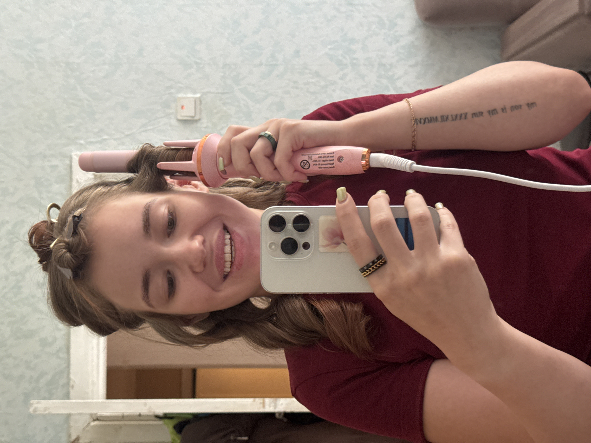 The image shows a young woman with brown hair, styled with hair rollers and a curling iron, looking at her phone. She is wearing a dark red top. The background appears to be an indoor setting, possibly a bedroom or bathroom, with a light blue patterned wall. A doorway with a partially visible interior is to her left. The woman is holding a smartphone in her left hand and a pink curling iron in her right hand. She has a green ring on her right ring finger and a black and gold ring on her left ring finger. A gold bracelet is visible on her right wrist. A tattoo is present on her right forearm. The overall scene suggests the woman is in the process of styling her hair. There are no specific indicators in the image that place it in Chaikovsky, Russia.