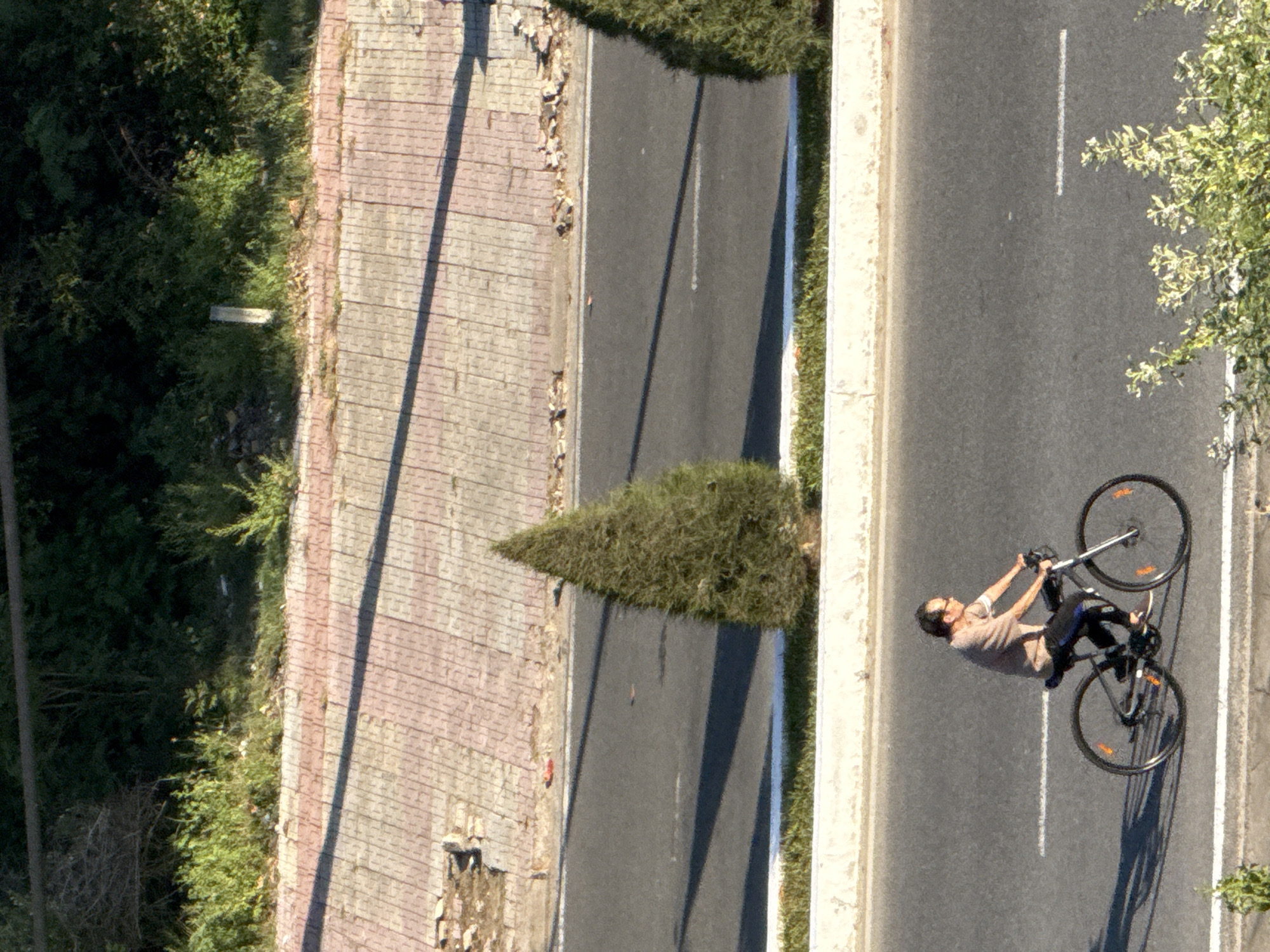 A person rides a bicycle on an asphalt road. The person is wearing a light brown shirt and dark pants. The bicycle is dark colored with orange reflectors on the wheels. The road is lined with a concrete barrier and a strip of grass. Behind the barrier, there is a tiled walkway with a shadow cast across it. Further back, there is a line of trees and vegetation. The scene appears to be outdoors in a city.