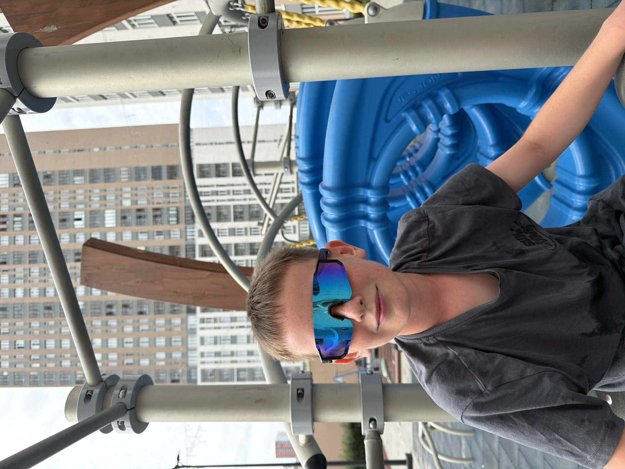 An outdoor, eye-level, medium shot shows a young Caucasian boy wearing a dark gray t-shirt and mirrored sunglasses with blue and purple lenses. He is smiling slightly as he looks at the camera. The boy is positioned in the foreground, seated in a playground structure with a large, bright blue plastic element behind him. The structure appears to be made of metal poles and supports, some painted a light beige. In the background, a cluster of tall, modern apartment buildings are visible under a cloudy sky. The architecture suggests an urban environment. The overall impression is that of a candid photograph taken on a day out at a playground in a residential area of Yekaterinburg, Russia.