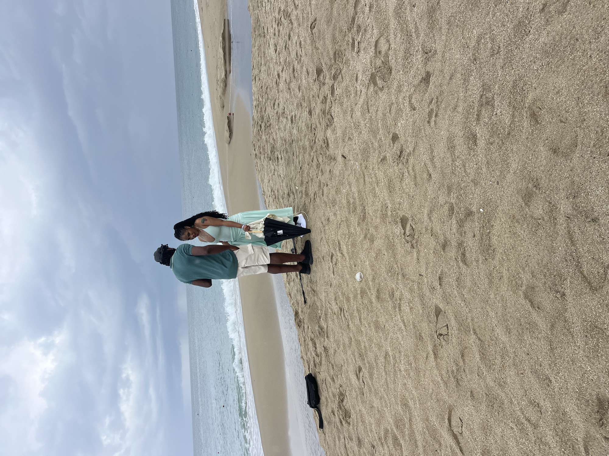 A couple stands on a sandy beach in Alanya, Turkey. The man, wearing a green shirt and khaki shorts, faces the ocean. The woman, in a light green dress, stands slightly behind him. An umbrella and a black bag sit on the sand near them. The ocean is visible in the background with waves breaking on the shore, and the sky is overcast.
