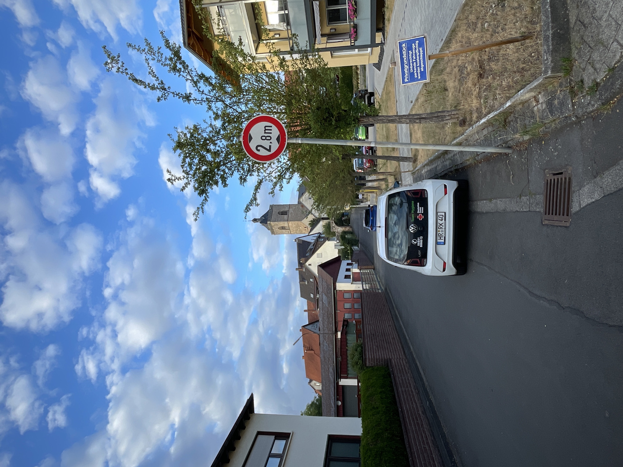 The image captures a street scene in Neukirchen, Germany. In the foreground, a white Volkswagen Up! is parked on the right side of the asphalt road, facing away from the viewer. The car has a license plate reading "HR RK 40" and displays stickers related to "up!" and "Team Hygiene" with a German Red Cross logo. A round traffic sign indicating a height limit of 2.8 meters is prominently placed on the right side of the road, near the car.
The street is lined with trees and houses. On the left, a white house with a grey-tiled roof and a dark-framed window with a closed roller blind is visible. A low brick wall topped with a hedge separates the property from the street. Further down the street, various residential buildings with red and grey roofs are visible. In the distance, a church with a prominent tower stands against a blue sky filled with white, fluffy clouds.
To the right of the road, beyond the trees and traffic sign, is a paved area and a patch of dry grass with a blue sign on a wooden post that reads "Privatgrundstück, Unberechtigt parkende Fahrzeuge werden kostenpflichtig abgeschleppt!" (Private property, Unauthorized parked vehicles will be towed at the owner's expense!). A modern apartment building with balconies and planters is also visible on the right. The overall atmosphere suggests a quiet, suburban or semi-urban environment.