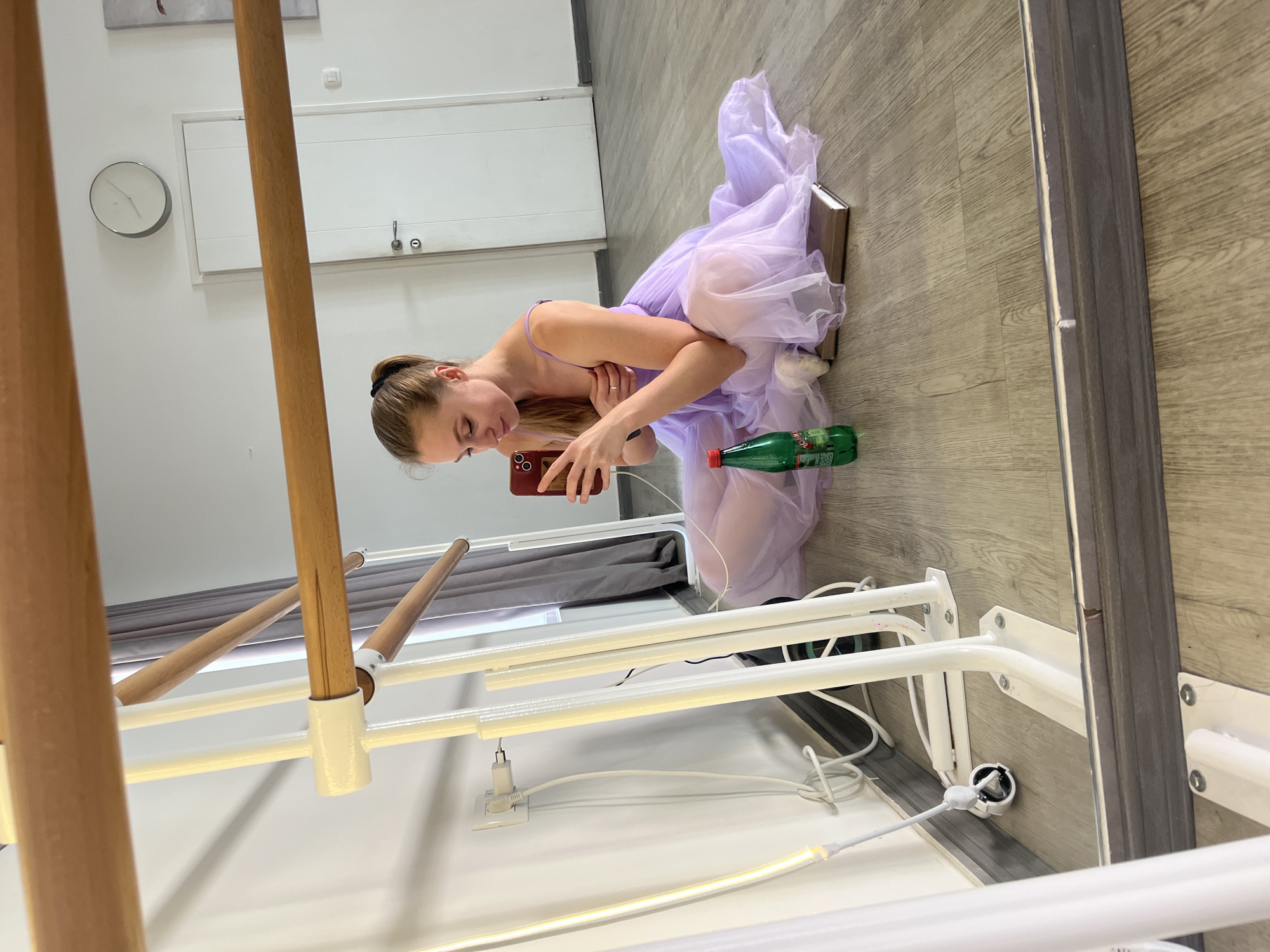 A young woman sits on the floor wearing a lavender tutu and holding a smartphone. A green glass bottle lies next to her. She is sitting on a closed book. The floor is light-colored wood. A ballet barre is in the foreground. The scene is indoors. A mirror is in the background. A wall with a clock and a small door is in the background. There is no indication of the location of Budva, Montenegro.