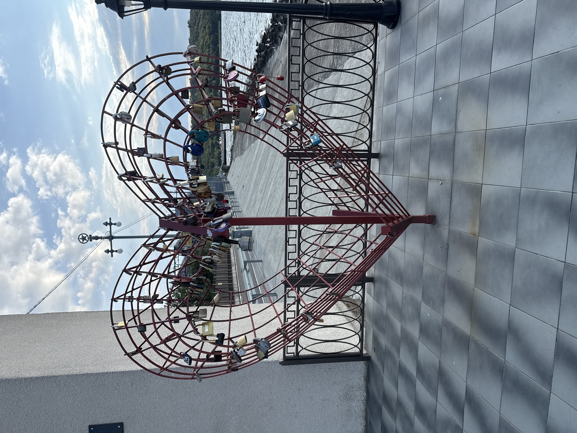 A large, red metal heart sculpture, adorned with numerous padlocks of various sizes and colors, stands prominently on a paved walkway. The heart sculpture is the central focus, with its intricate wireframe construction visible, supporting the attached locks. Behind the sculpture, a paved walkway leads towards a body of water, likely a river or lake, with a tree-lined shore visible in the distance. A black metal railing borders the walkway in front of the water. To the left of the heart sculpture, a textured, light-colored wall occupies the foreground, and a black lamppost stands to the right. The sky above is partly cloudy, with scattered white clouds against a pale blue backdrop. The scene suggests a public waterfront area, possibly a promenade or park, in Korzhevo, Moldova, a location known for its scenic riverbanks. No people are clearly visible in the image, and no discernible actions or interactions are taking place.