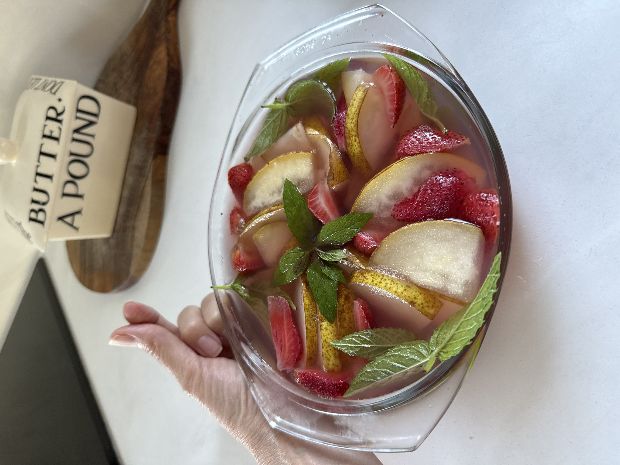 A person's hand, with fingers curved to form a "finger heart" gesture, is visible in the upper left quadrant of the image. The hand is positioned above a clear glass bowl filled with a pinkish liquid. The bowl contains slices of pear, strawberries, and mint leaves. In the background, a wooden cutting board supports a white ceramic butter dish with the words "BUTTER. A POUND." printed on it. The surface beneath the bowl and butter dish appears to be a white countertop. The scene is indoors, on a bright, well-lit day. The location is not specified as Steklyannyi, Russia.