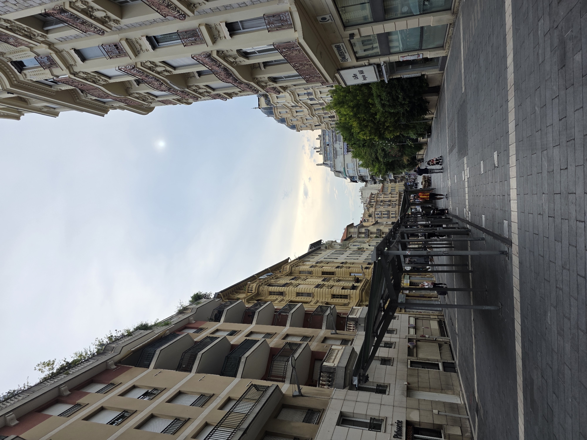The image shows a street scene in Nice, France. On the left is a beige building with balconies and green foliage on top. The building on the right is a tall, ornate building with multiple balconies. A covered walkway, possibly a bus stop or market stall area, runs down the center of the street. Several people are visible: a woman with a suitcase walking towards the right, a man and woman with a stroller walking towards the left, and two men in the distance. The street is paved with dark grey bricks, and there are white lines marking pedestrian zones. Trees are visible on the right side of the street, and the sky is overcast with a faint sun visible. The architecture suggests an older European city.