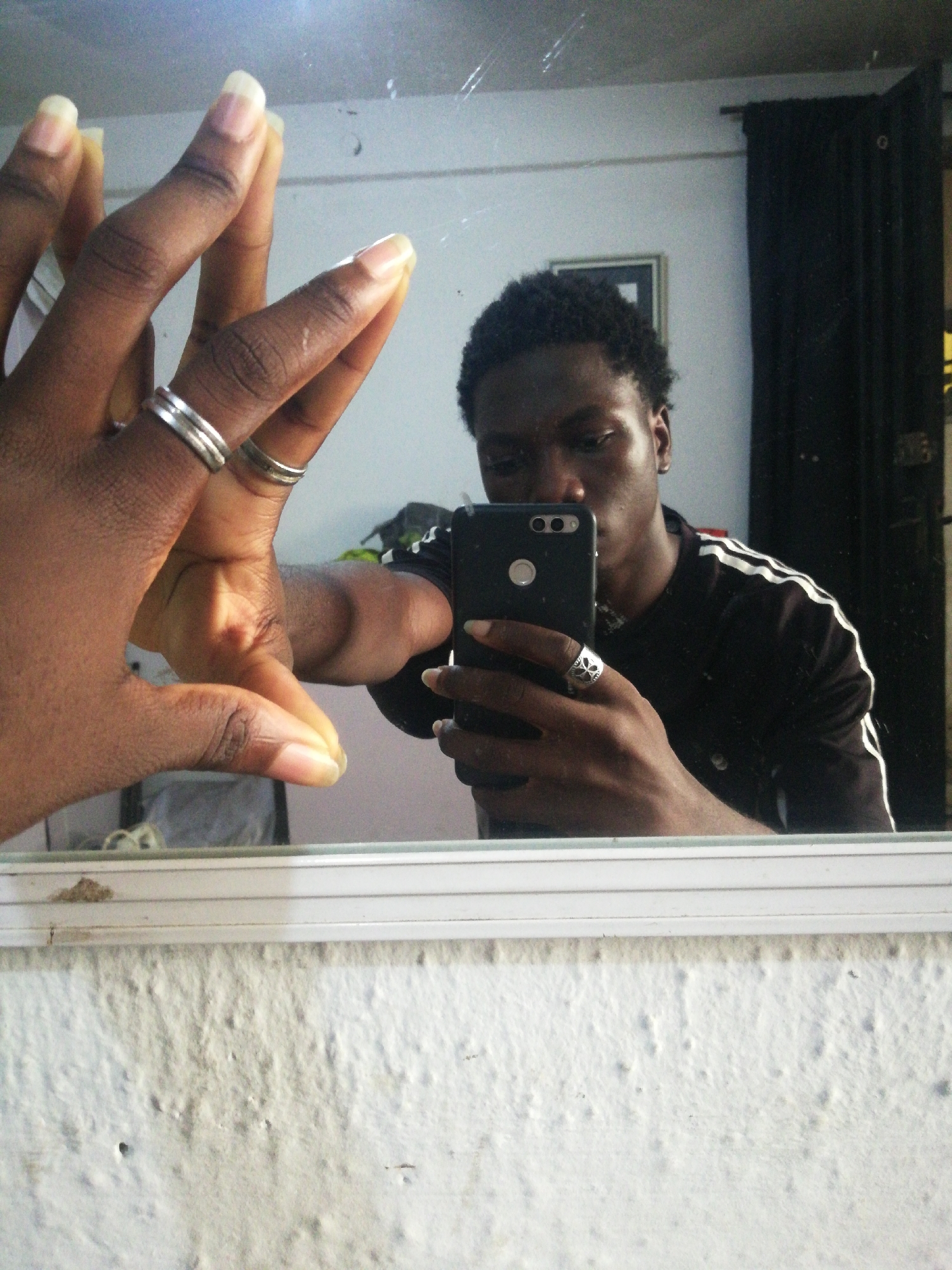 A person is taking a selfie in a mirror. The person is dark-skinned, with short, dark hair, and wearing a black shirt with white stripes on the sleeves. They are holding a black smartphone in their right hand and there is a ring on the fourth finger. The mirror shows the person's hand with two rings on the left. The background of the mirror reflection shows a wall with a picture frame and a black curtain. The wall in front of the mirror has a textured surface. The location is Lagos, Nigeria.