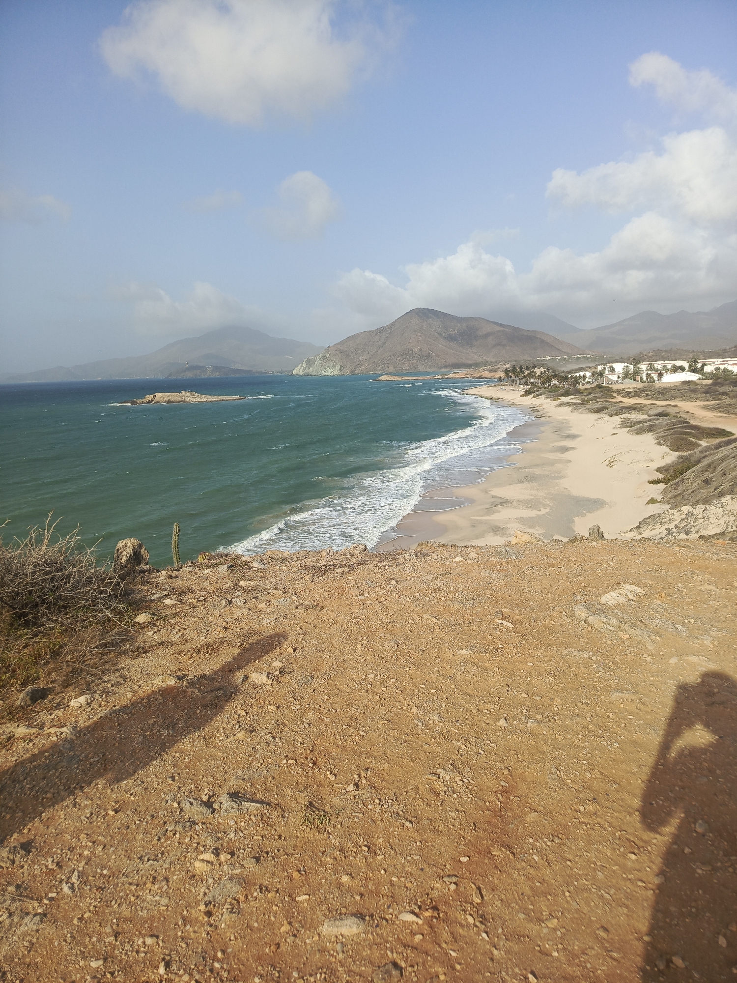 The image shows a coastal scene in Altagracia, Venezuela. The foreground consists of a brown, rocky cliff face. In the middle ground, there is a beach with light-colored sand and ocean waves meeting the shore. The water transitions from a teal color in the distance to foamy white near the beach. Beyond the beach, there are buildings and vegetation. In the background, there are mountains and a slightly hazy sky with scattered clouds. There is a shadow of a person taking the photo in the lower right. A small cactus plant is visible on the cliff in the middle-left.
