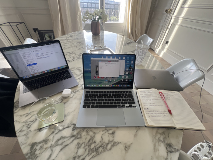 The image depicts a well-lit interior scene, likely a modern apartment in Paris, France, identifiable by the Haussmannian-style balcony railing visible through the window. The primary focus is an oval white marble table serving as a workspace.
On the table, three Apple MacBook laptops are present. One space gray MacBook on the left is open, displaying an email application's inbox. Next to it is a white Apple AirPods case and a clear glass of water on a light green coaster. A second, silver MacBook is open in the center, displaying a macOS desktop with various application icons and a prominent white modal window overlaying the screen. To the right of this laptop is an open notebook filled with handwritten text in black ink, containing a red pen with white polka dots resting within its pages. A third, silver MacBook is closed and positioned behind the central laptop and notebook. A white disposable cup with a lid and straw is also on the table near the closed laptop.
The background reveals a brightly lit room with large windows draped in translucent white curtains. Strong sunlight casts distinct shadows across the white paneled walls and the light-colored wooden herringbone floor. Through the windows, a glimpse of the classic Parisian architectural style, including a metal balcony railing and distant trees, is visible. Two modern, light-colored chairs are partially tucked under the marble table. A brown vase containing white flowers is positioned on the table directly in front of the window. A dark, geometric metal structure is partially visible in the far left background, and a golden doorknob is discernible on a paneled door on the right. No individuals are present in the scene.