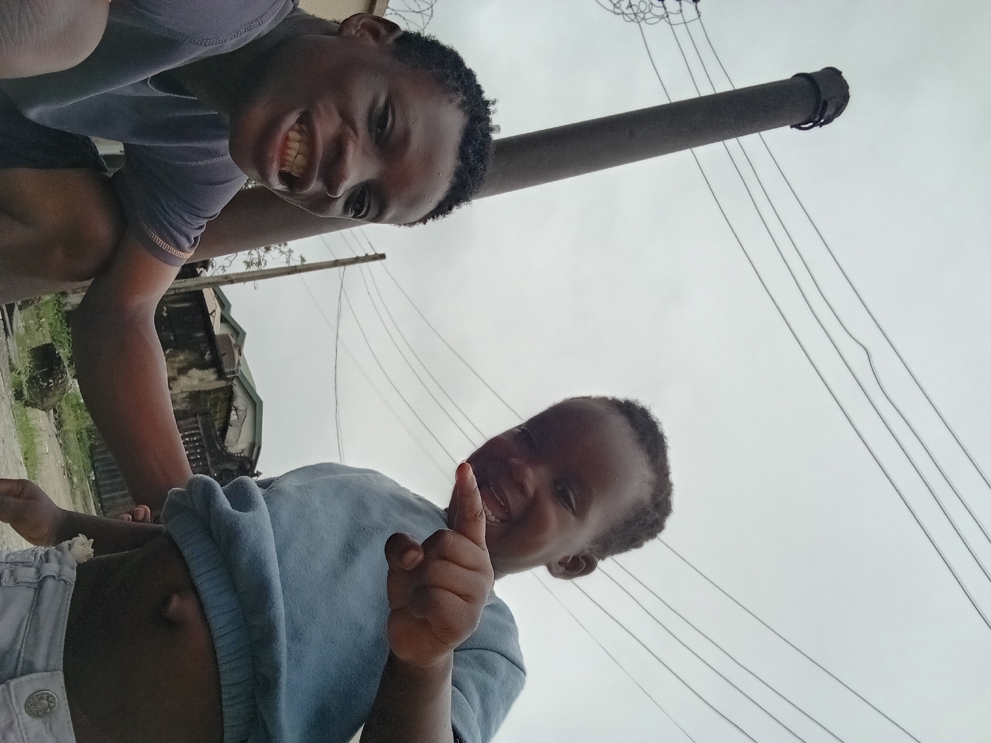 A low-angle outdoor photograph shows two young Black boys in front of a pole and power lines against an overcast sky. The boy on the left, closer to the camera, is wearing a dark blue t-shirt. He is crouched and smiling broadly, with his mouth open and teeth showing. His left arm is bent, with his hand resting on his knee. The boy on the right is pointing his right index finger forward, and smiling. He is wearing a light blue sweatshirt and light-colored shorts. The background shows a fence, buildings, and vegetation.