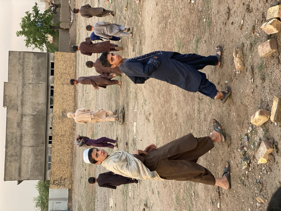 The image depicts an outdoor scene in Kandahar, Afghanistan, featuring approximately eleven young males on unpaved, dry, earthen ground. The sky is overcast, indicating diffused lighting conditions.
In the foreground, two young males walk towards the left, looking back towards the camera. The male on the left wears a brown shalwar kameez, a light-colored striped shawl draped over his shoulders, and a white skullcap (taqiyah). He has open-toed sandals. The male on the right wears a dark blue shalwar kameez with embroidered details on the chest and sleeves, also with open-toed sandals. Several broken reddish-brown bricks are scattered on the ground directly in front of them.
In the mid-ground, a cluster of young males, all wearing traditional shalwar kameez in various shades of brown, beige, purple, and grey, are either walking away from the camera or standing. One individual in beige clothing stands with his back to the viewer, positioned next to a large stone. Another individual in purple clothing is carrying something wrapped in fabric over his shoulder. The ground is uneven, dusty, and littered with small stones and debris.
The background features a two-story, unrendered concrete building with several rectangular window openings. To its left, a corrugated metal fence is visible, and below the concrete building, a light-brown brick wall extends across the mid-background. A single green-leafed tree stands to the right of the concrete building, behind which another lower concrete structure is visible.