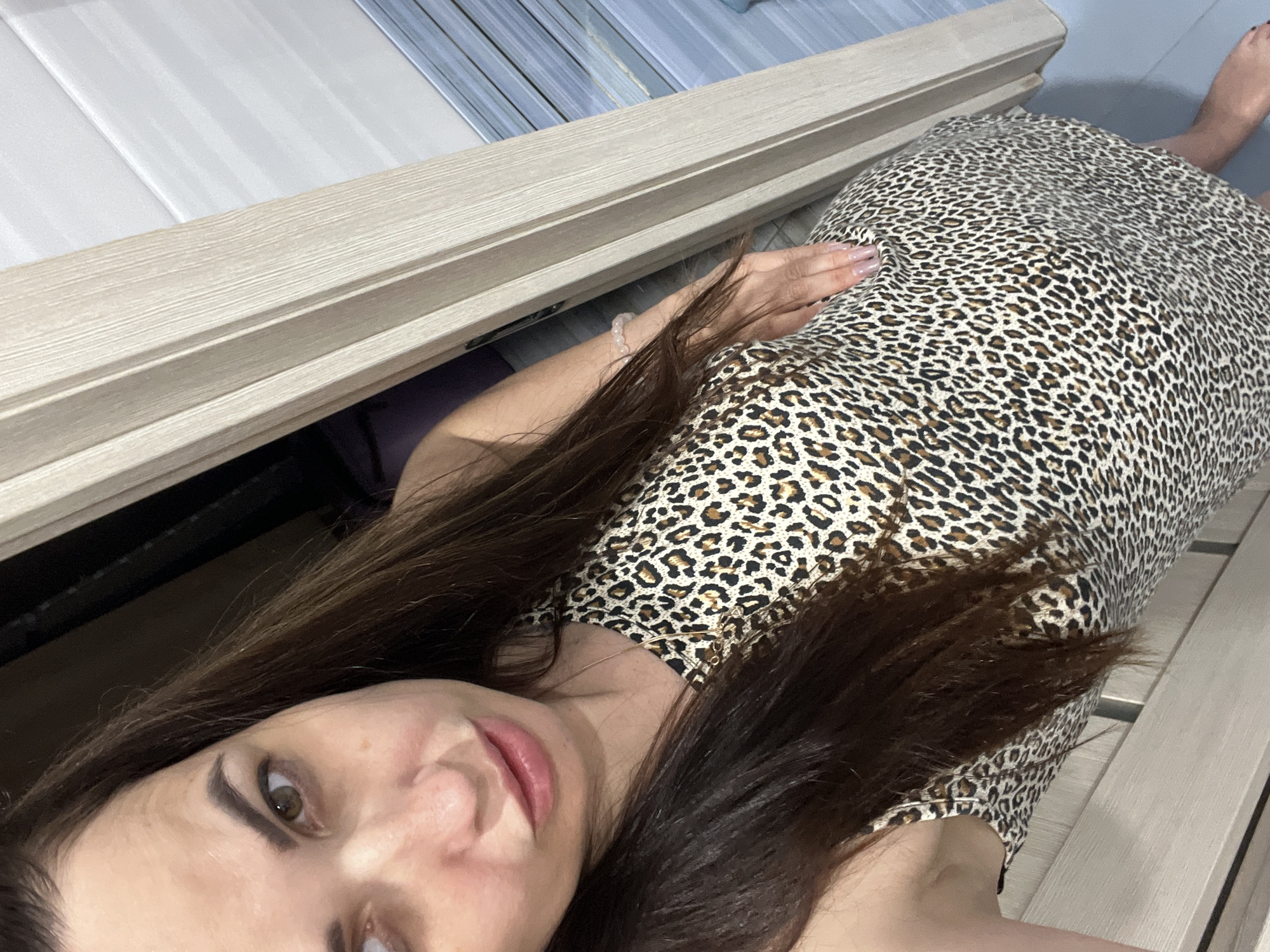 A woman is standing in a doorway, wearing a leopard-print dress. She is looking at the camera and her left hand is resting on her hip. The background shows a light-colored doorframe and a tiled wall.
