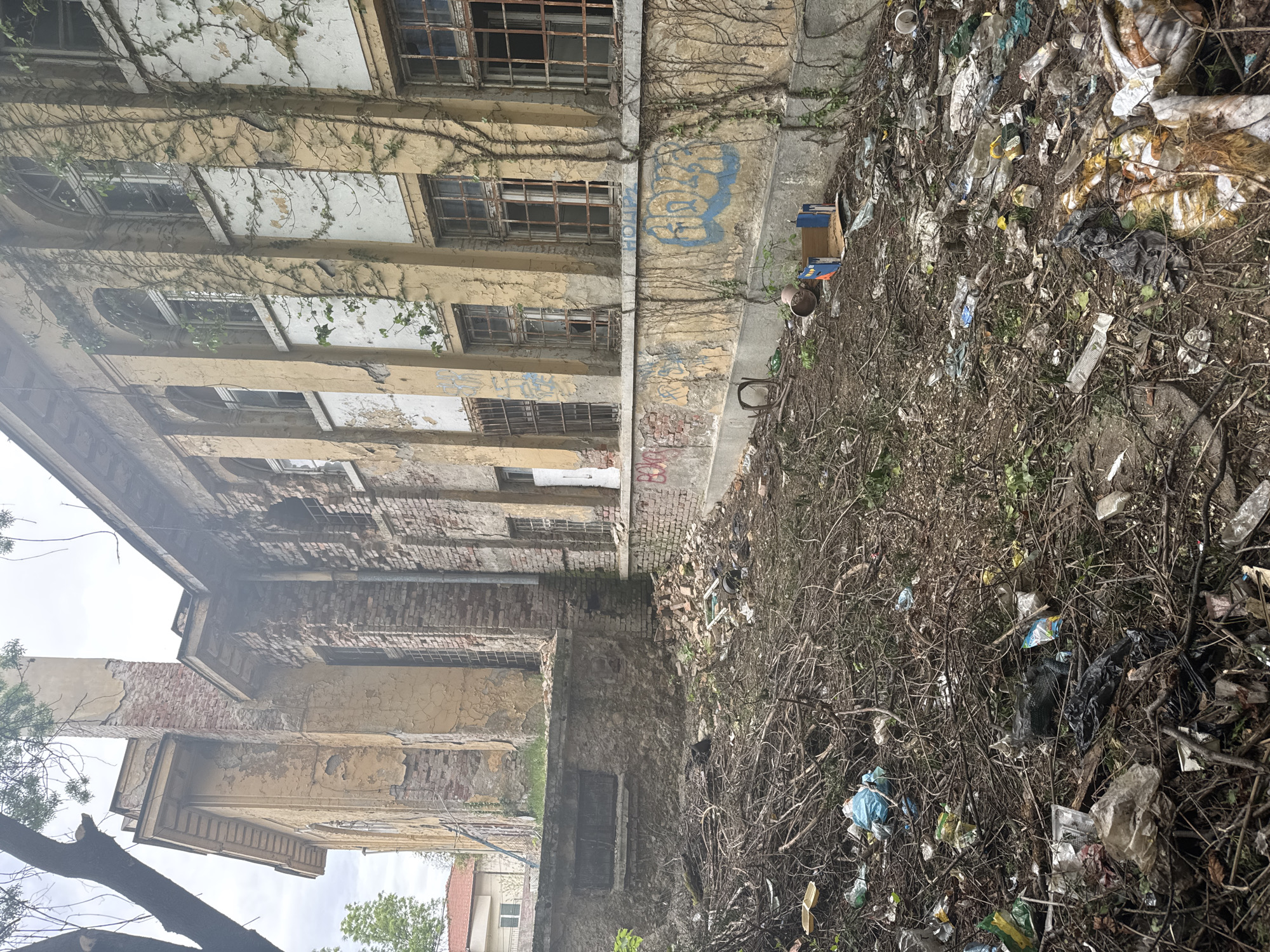 The image shows the exterior of a dilapidated building in Sofia, Bulgaria, possibly abandoned. The architecture features multiple stories with deteriorating facades, weathered paint, and numerous windows, some boarded up or broken. Vines climb the building's walls. The ground level is obscured by a dense layer of debris including litter, discarded objects, and dead branches. The overall impression is one of urban decay.