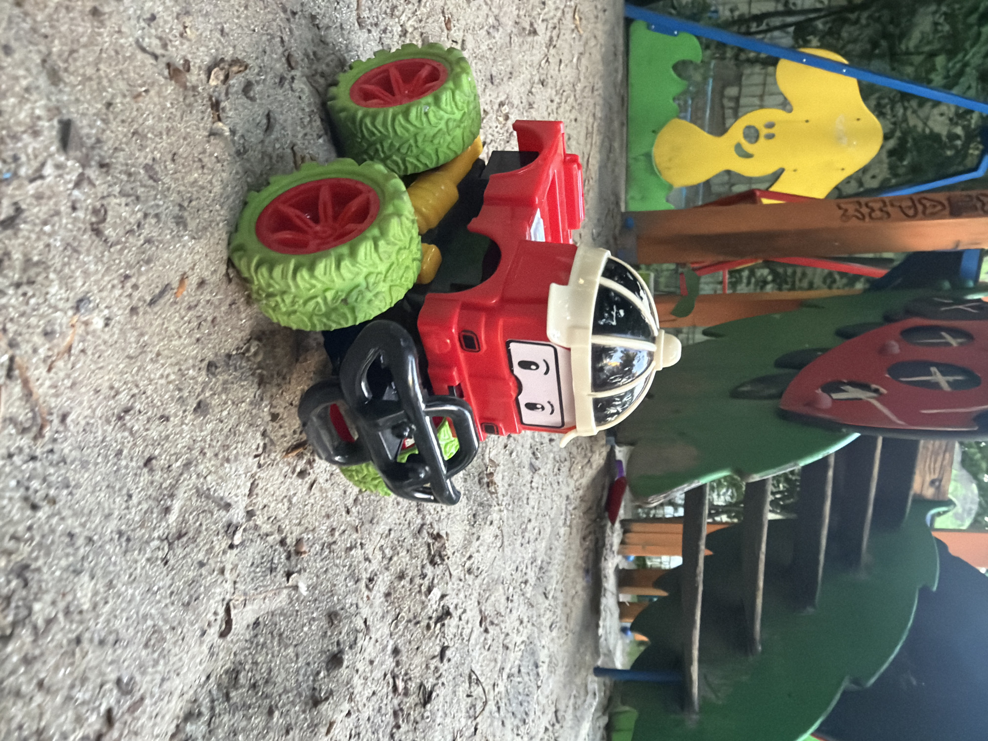 A toy monster truck, primarily red with a white helmet-like structure on top, sits on a sand-covered surface. The truck has large green tires with red rims and a black bumper. In the background, a portion of a playground structure is visible, including green and red elements, wooden stairs, and a yellow cutout of a smiling face. The scene appears to be outdoors, possibly in a park or playground, given the sand and playground equipment.