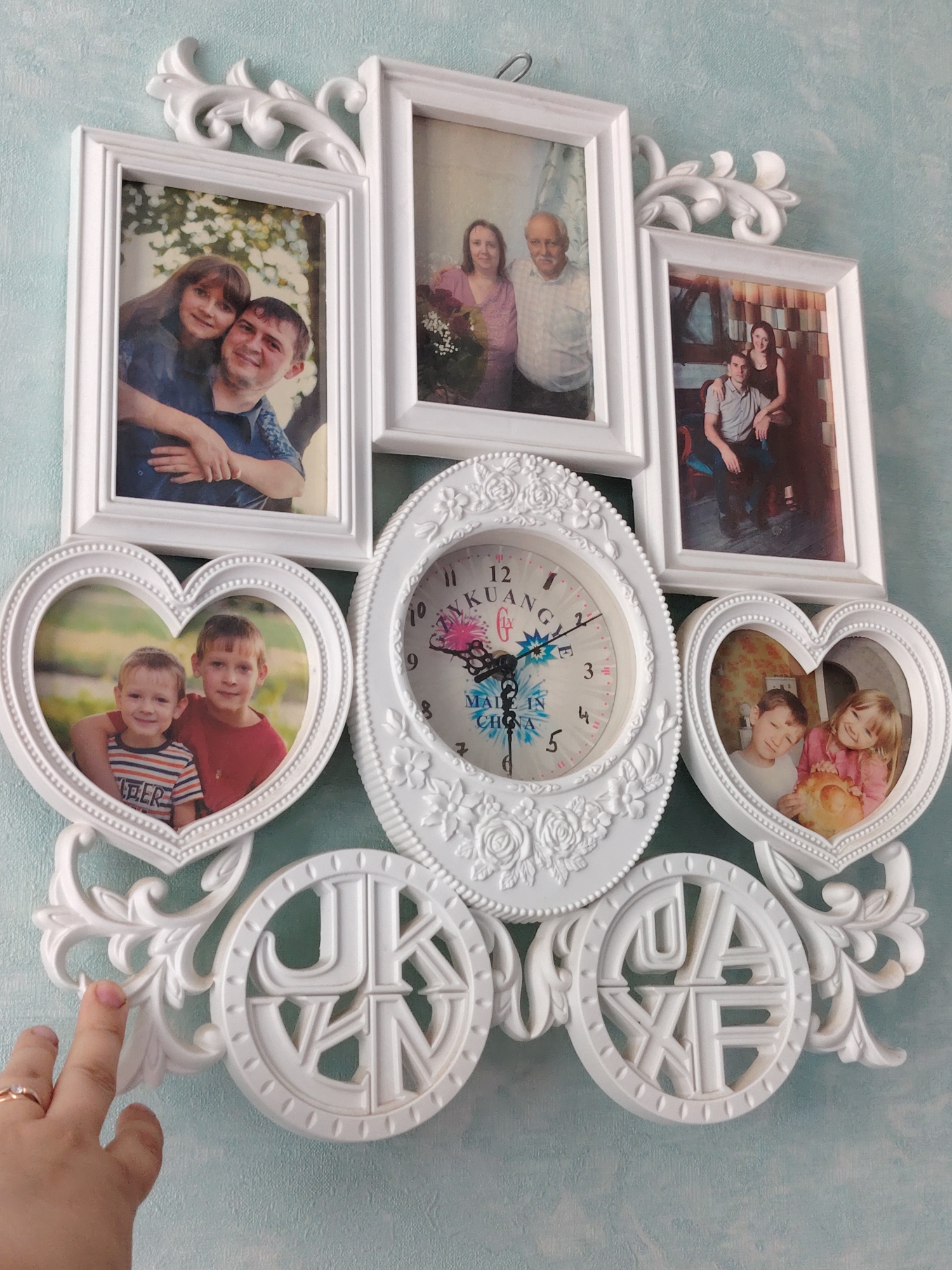 This is a close-up, eye-level shot of a white, ornate wall clock with multiple photo frames, mounted on a textured, pale blue wall. The clock is centrally located, featuring an oval-shaped face with a floral decorative border. The clock face itself has Roman numerals and hands, indicating a time around 11:45. The clock's design includes the words "MADE IN CHINA."
Surrounding the clock are five white photo frames, arranged in a staggered, symmetrical pattern. The top row has two rectangular frames and one larger rectangular frame in the center. The bottom row has two heart-shaped frames, one on each side of the clock. Each frame displays a photograph:
* **Top left frame:** A couple is pictured, with the woman leaning on the man's shoulder. They are smiling and appear to be outdoors.
* **Top center frame:** An older couple is photographed, the man with a mustache. The woman is wearing a pink top, and the man is in a white, patterned shirt. There are flowers in the woman's hand.
* **Top right frame:** A couple is seated, with the man arm around the woman.
* **Bottom left heart frame:** Two young boys are pictured, one with a striped shirt and the other in a red shirt. They are smiling and positioned closely together.
* **Bottom right heart frame:** A young boy and girl are photographed. The girl is holding a round, light-brown object, possibly bread, and both are smiling.
Below the heart-shaped frames, two circular decorative elements with intricate cutouts of letters are attached. The left one displays the letters "JK" on the top half and "VN" on the bottom half, within a circular border. The right decorative element shows "UA" on the top and "XF" on the bottom.
A person's hand with a ring on the ring finger is visible in the lower left corner, touching a decorative scrollwork element of the clock's frame. The background is a textured, pale blue wall, common in interior settings. The overall aesthetic suggests a decorative wall piece designed to display family photographs and tell time. There is no information in the image to indicate the location in the city of Volnoye, Russia.