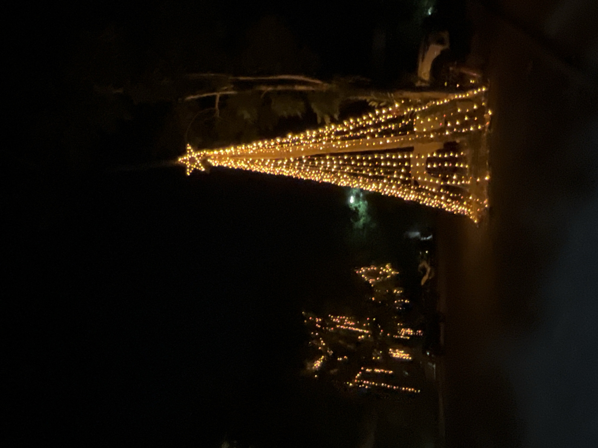 At night, a tall, cone-shaped Christmas tree decorated with warm yellow lights stands in the foreground. The lights are arranged in a dense pattern, forming the outline of the tree and culminating in a star at its apex. To the left of the main tree, another cluster of illuminated trees or decorations displays similar warm yellow and orange lights, creating a festive atmosphere. In the background, dark trees are visible, and to the right of the main tree, a portion of a white vehicle is partially obscured by the tree's lights. The scene appears to be set in an outdoor area, possibly a street or park, within the city of Ban Rak, Thailand, given the context provided. The ground in the foreground is dark and indistinct.