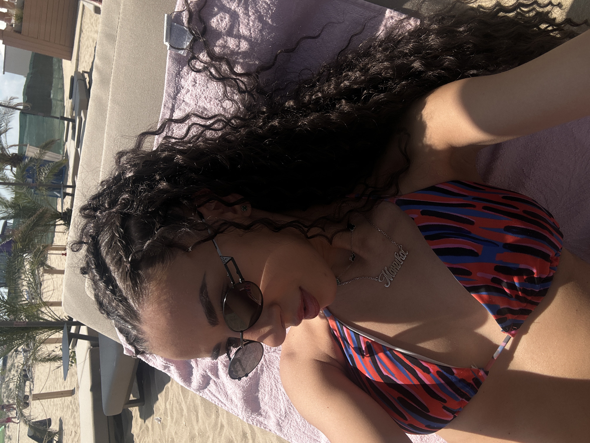 A young woman is lying on a beach lounge chair on a sandy beach. She is wearing a colorful bikini and sunglasses. Her dark, curly hair is partially braided and spills over her shoulders. A silver necklace with a nameplate is visible on her chest. The background shows palm trees and more lounge chairs, suggesting a resort or beach club. The lighting indicates it is a sunny day. This scene is situated on the beach in Nesebar, Bulgaria.