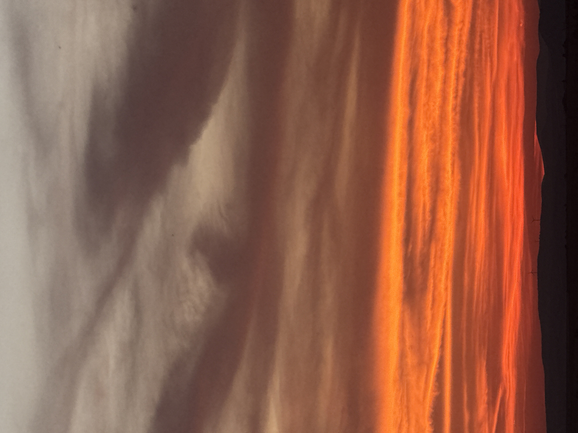 The image depicts a vibrant sunset over the city of Burgas, Bulgaria. The sky is filled with dramatic clouds, transitioning from a pale grey and muted orange at the top to intense fiery orange and red hues towards the horizon. These colors are further accentuated by horizontal bands of lighter clouds, creating a layered effect. In the foreground, silhouetted against the glowing sky, is a dark, undulating landscape, representing the hills or coastline of Burgas. A few wind turbines are faintly visible on the horizon, suggesting a more rural or industrial edge of the city. The overall scene is one of natural beauty and atmospheric grandeur, captured during the twilight hours.