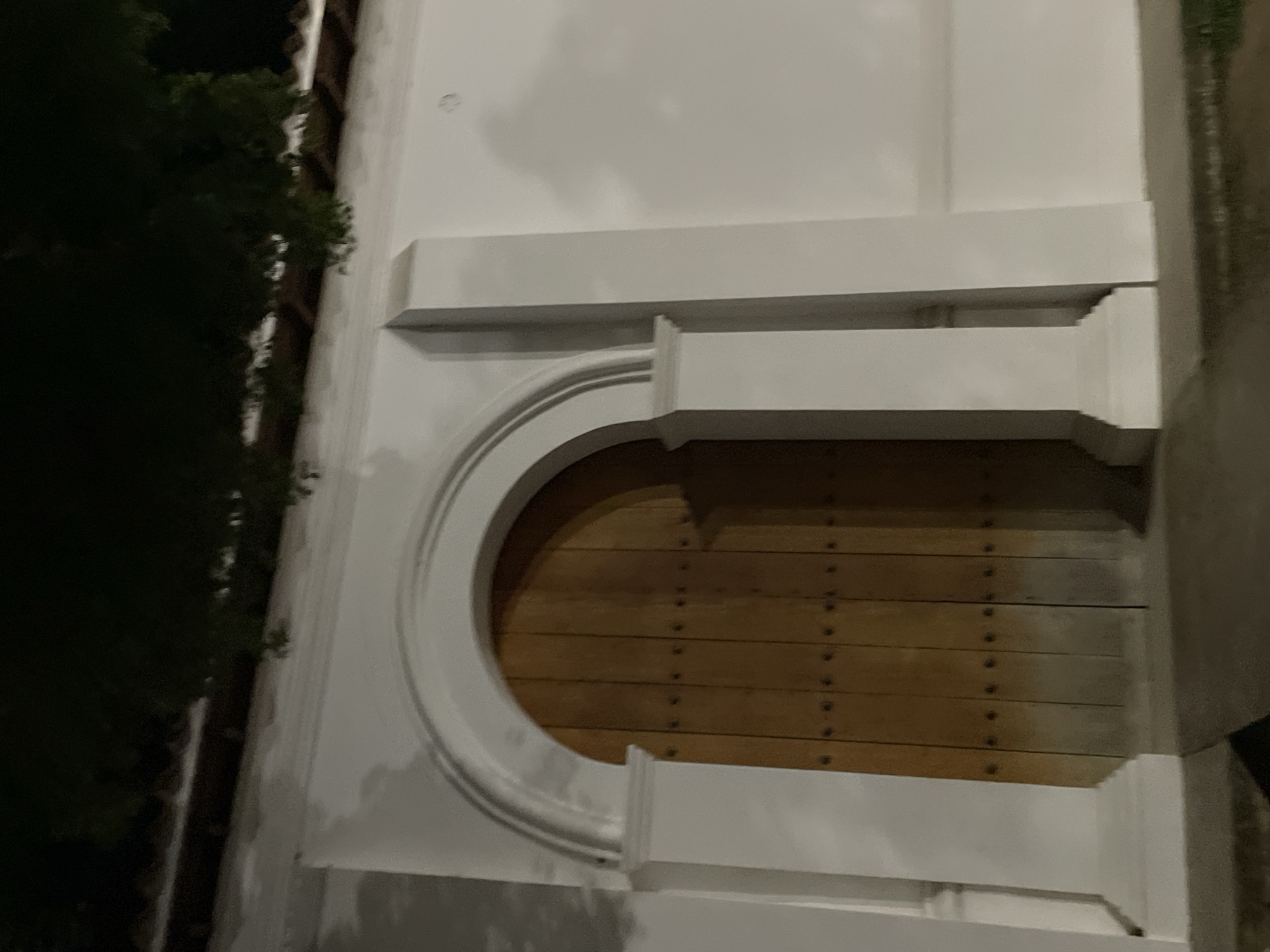 The image shows a large wooden double door set within an arched, white-stone entryway. The door has a light brown, weathered finish and is adorned with rows of dark, circular studs. The entryway features two substantial white pillars flanking the door, topped with decorative elements. The wall surrounding the entryway is also white. Above the entryway, a dark green canopy of leaves from unseen trees extends over the scene, casting shadows. The overall impression is of an old, possibly historical, building entrance. The image is taken at night, with artificial lighting illuminating the white surfaces and creating soft shadows. The scene is devoid of people and any visible activity. The location is identified as the city of Santa Ana, Venezuela.