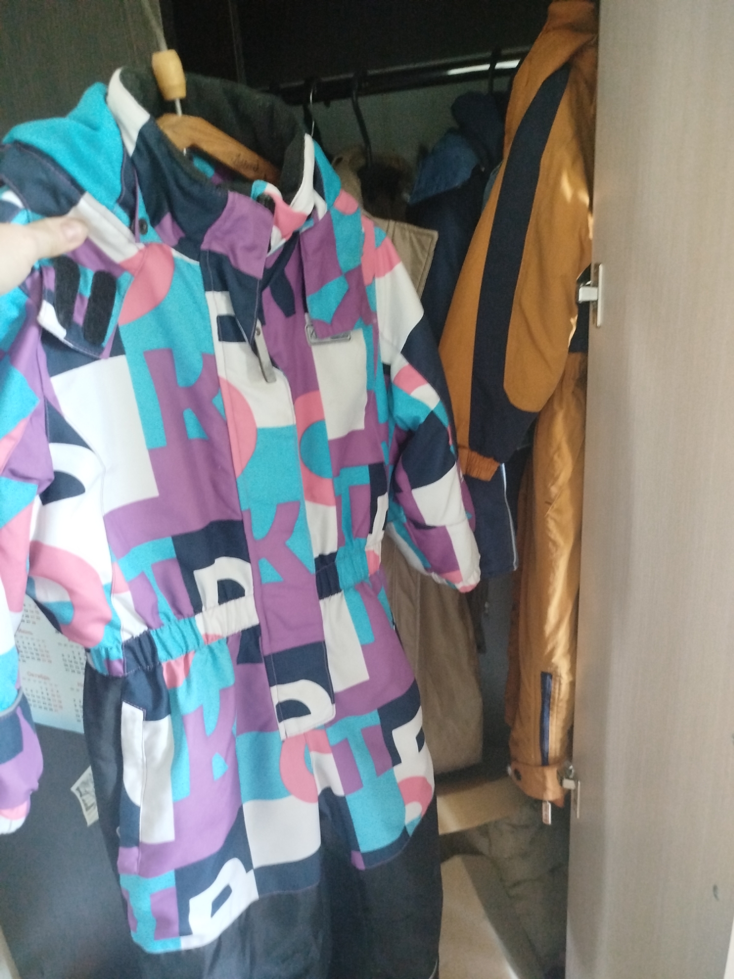 An interior shot shows a colorful ski suit hanging on a wooden hanger, positioned on the left side of the frame. The suit is predominantly purple, turquoise, white, and pink, with a geometric pattern of large shapes and letters. A human hand is visible on the left, appearing to adjust the suit. Behind the ski suit, other jackets are hanging in a closet. To the right, a door with a silver handle is partially visible. The background is dark and indistinct. The scene is a closet in Zavodoukovsk, Russia.