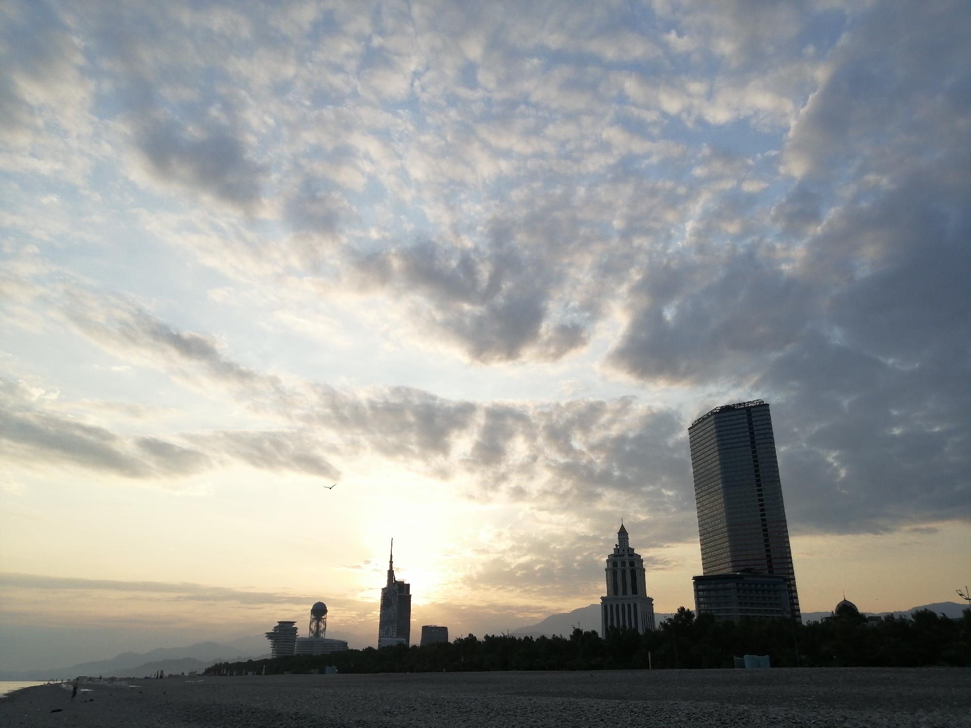 The image shows a wide shot of the Batumi coastline at sunset. The sky is filled with dramatic clouds, tinged with the warm hues of the setting sun. In the foreground, a pebble beach stretches out, with a few distant figures visible on the sand.
Silhouetted against the sky are several modern buildings, characteristic of Batumi's urban landscape. To the left, a spherical structure atop a building is visible. Further to the right, a tall, slender skyscraper with a glass facade leans slightly, while a more ornate, tower-like building with a pointed top stands nearby. The buildings are nestled behind a line of dark trees. In the far distance, a range of mountains forms a hazy backdrop. A lone bird is captured in flight against the vast expanse of the sky. The overall scene conveys a sense of calm and the beauty of a coastal city at dusk.