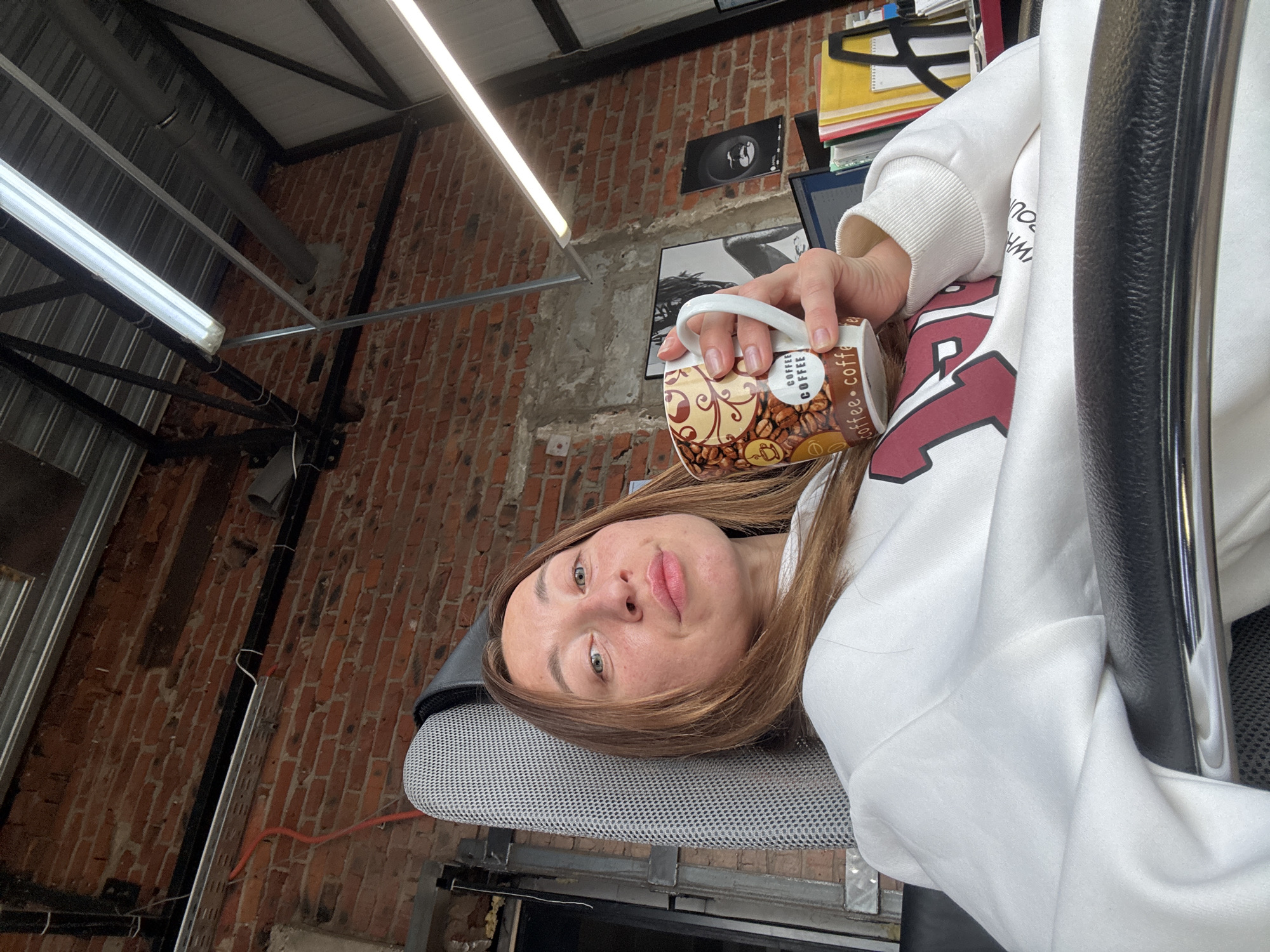 The image shows a woman sitting in an office chair, holding a coffee mug with coffee bean designs and the word "COFFEE" printed on it. She is wearing a white sweatshirt with red lettering on the front. The office has a brick wall, and the ceiling is exposed with metal beams and fluorescent lights. There is a monitor and other office items in the background, including a stack of papers in a yellow folder. The chair is gray with a black frame. The photo appears to be taken at an angle from above. The specific location of this scene is not mentioned.