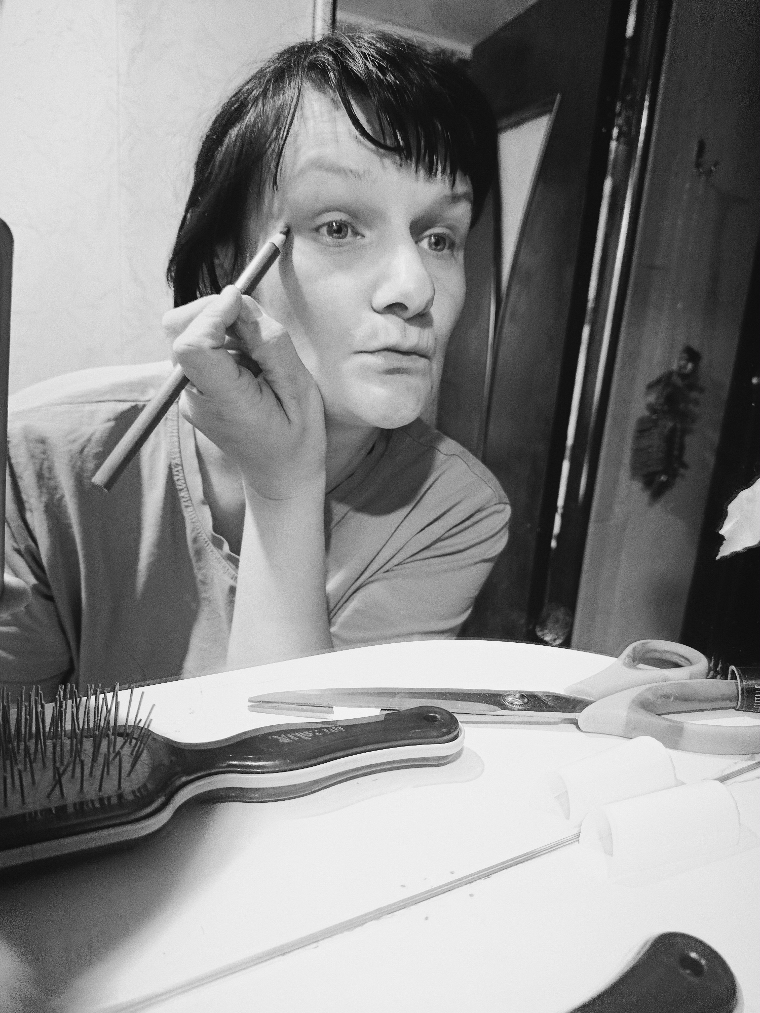 The black and white image captures a person with short, dark hair applying makeup, likely eyeliner, to their right eye using a pencil. They are looking intently into what appears to be a mirror, or directly at the camera lens, with a focused expression. Their left hand is resting near their chin, supporting their head or holding something out of frame. The person is wearing a light-colored t-shirt.
In the foreground, on a reflective white surface, several items are visible. To the left is a hairbrush with a black handle and numerous bristles, partially obscured. A partially legible brand name, "SCALPM...", can be seen on its handle. To the right of the hairbrush, a pair of scissors with distinct light-colored handles and metal blades lies flat. Further to the right are two white, cylindrical objects, possibly hair rollers, threaded onto a thin rod.
The background consists of a light-colored wall or surface on the left and a darker, vertically paneled structure, possibly a wooden door or cabinet, on the right. A faint, dark reflection or object appears on the dark background element. The overall lighting suggests an indoor setting, with illumination primarily coming from the front, highlighting the person's face and the foreground objects. The monochromatic tone gives the image a somewhat stark and timeless quality.