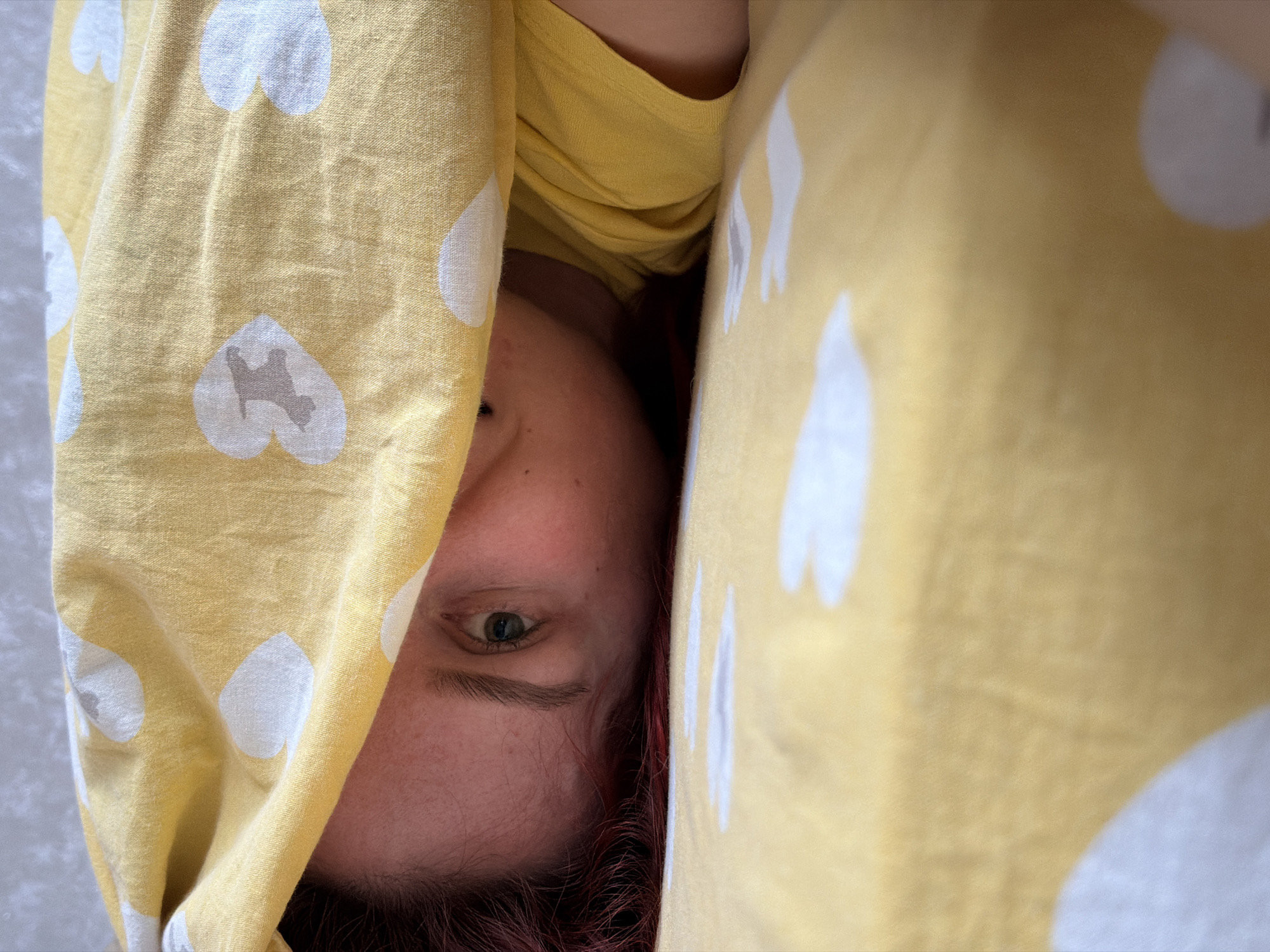A close-up, sideways shot depicts a person peeking out from under yellow bedding adorned with a pattern of white hearts. The subject's left eye, a portion of their forehead, and reddish-brown hair are visible, positioned on the right side of the frame and looking left. They appear to be wearing a yellow top that coordinates with the bedding. The visible eye holds a neutral, possibly watchful or curious expression. One of the white hearts on the fabric contains a small, light brown silhouette, resembling a stylized animal, possibly a dog. The background beyond the covers is an indistinct, light-colored, textured surface. The scene suggests an indoor setting, likely a bedroom in Chaikovsky, Russia. The activity is that of someone looking out from under covers, perhaps waking up or relaxing. There is no visible text within the image, and the soft lighting does not indicate a specific time of day.