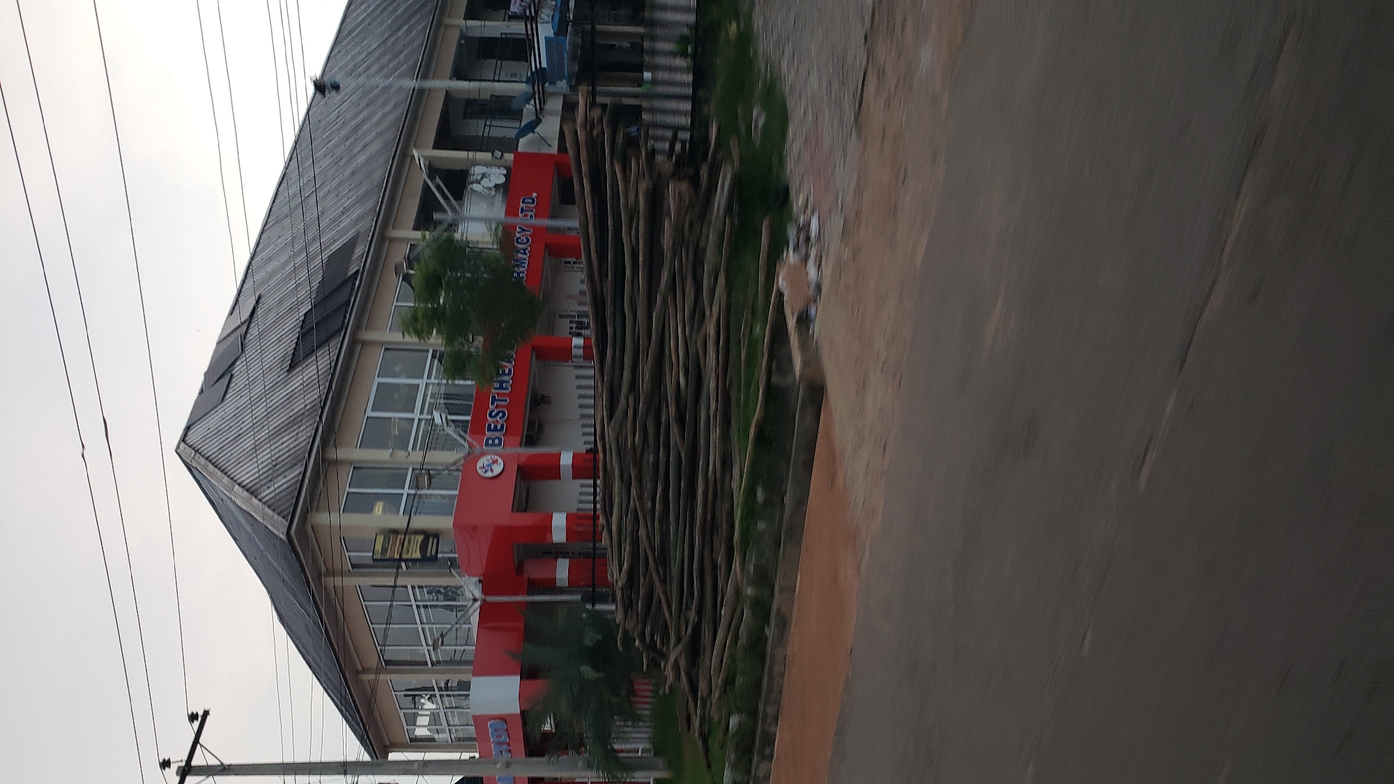 In Jalingo, Nigeria, a two-story building with a distinctive red and beige facade stands prominently. The building's roof is covered in corrugated metal sheeting, with some sections appearing to have solar panels installed. The upper floor features large glass windows, reflecting the overcast sky. A red awning with white trim extends across the lower level. Above the awning, the words "BESTHEALTH PHARMACY LTD." are visible in white lettering. A large pile of long, thin logs lies on the ground in front of the building, extending towards the street. The logs are stacked horizontally and appear to be in a natural, unworked state. The street in the foreground is paved with asphalt, and a dirt embankment is visible to the left. Utility poles and power lines are present in the background, suggesting an urban or semi-urban setting. The overall scene depicts a commercial establishment with construction or storage materials on display, possibly indicating ongoing development or a business related to lumber.