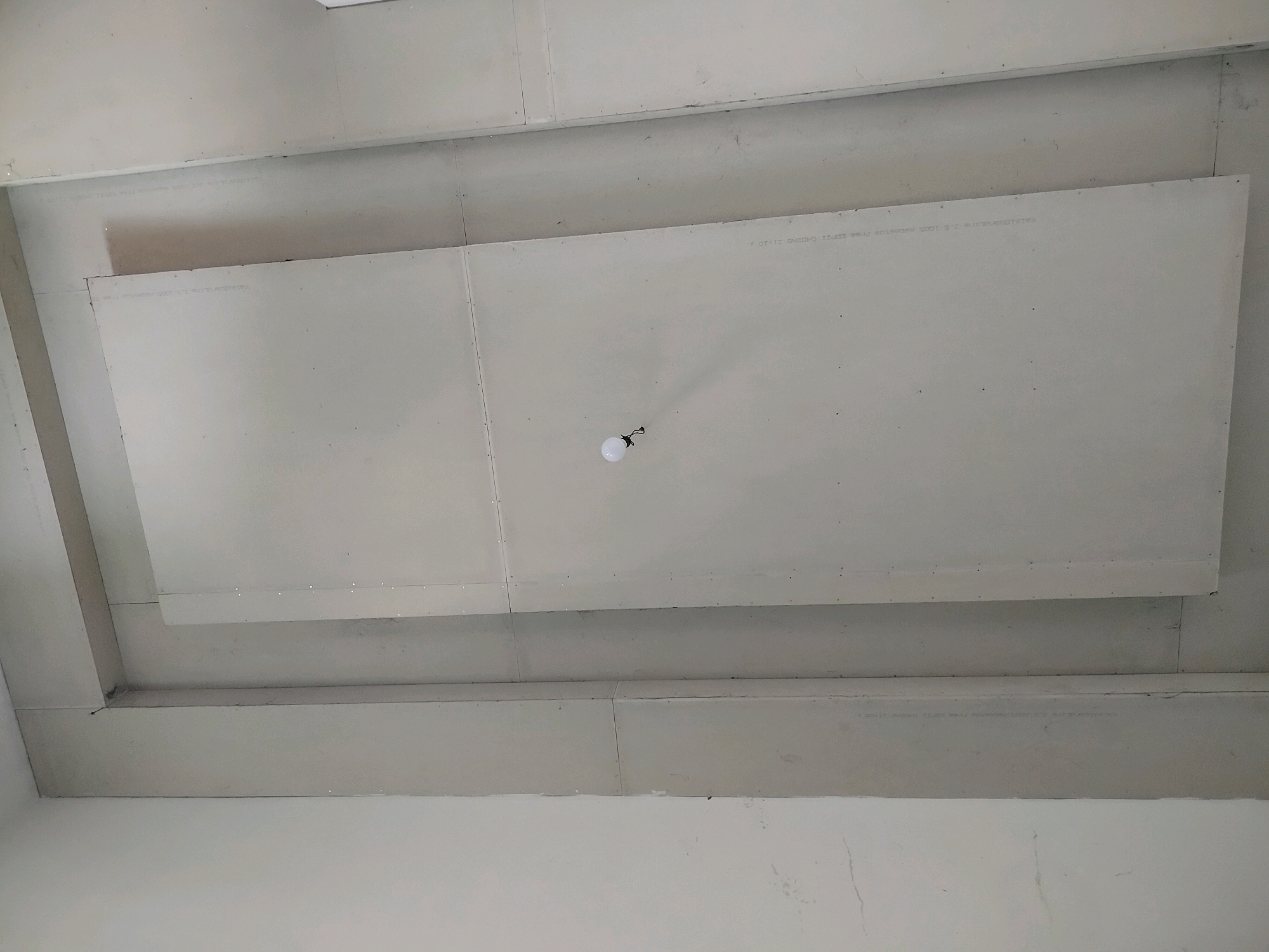 This image shows a section of a white, unfinished ceiling, likely in a residential or commercial building under construction. The ceiling is constructed from large, rectangular drywall panels, some of which are overlapping. There are visible seams and screw indentations on the panels. A single, bare lightbulb hangs from a wire in the center of the frame, illuminating the area below. The overall impression is one of a basic, functional ceiling structure. The image does not provide any information about the specific location in the city of Kroya, Indonesia, nor does it depict any people, actions, or interactions.