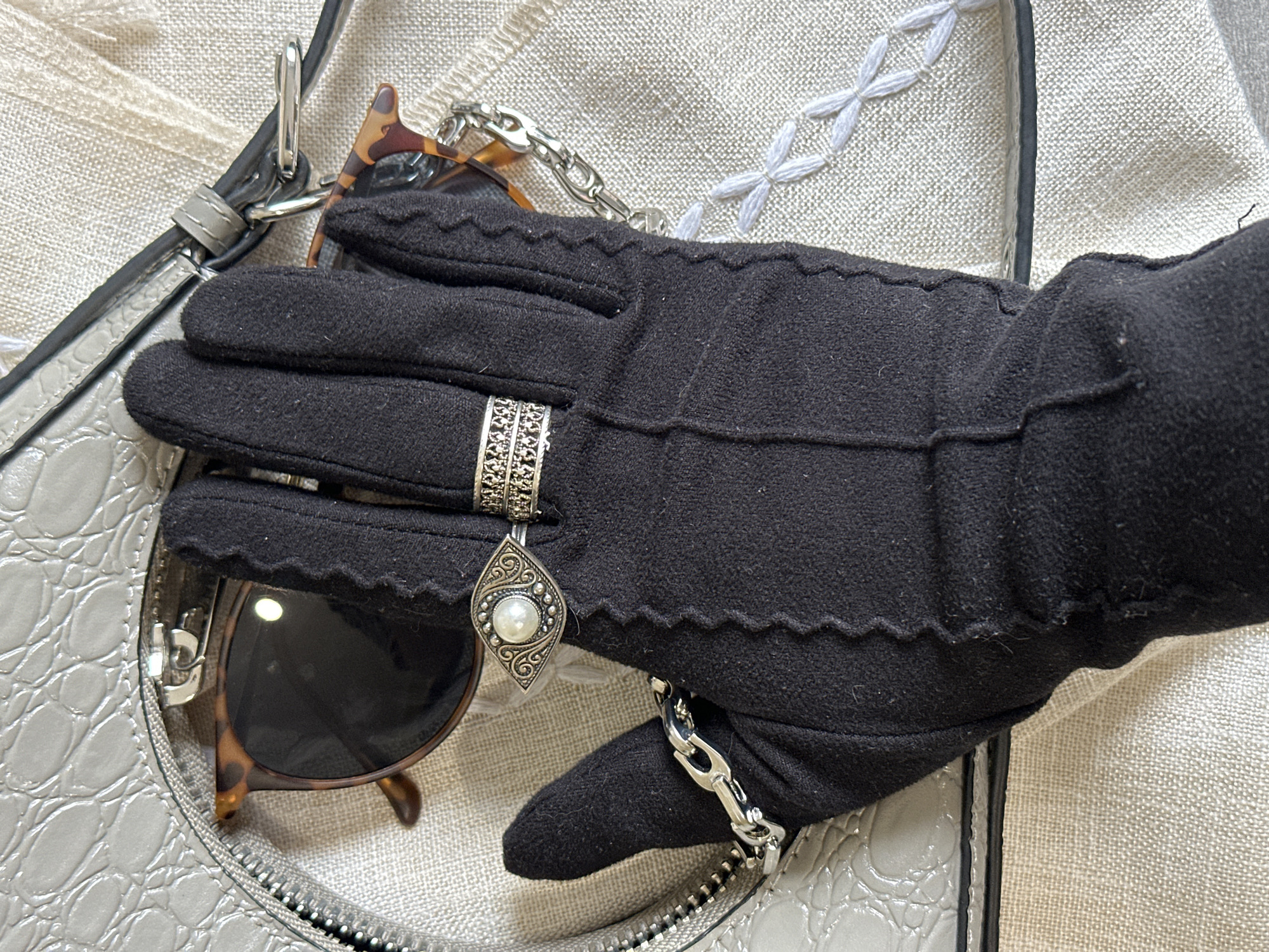 The image presents a close-up, high-angle flat lay showcasing a collection of fashion accessories arranged on a light beige or off-white textured fabric surface, possibly linen, featuring decorative white cross-stitch embroidery.
The central subject is a hand wearing a fitted black glove with scalloped edges and visible seam lines. On the glove's ring finger, an intricate silver-toned ring is worn, featuring a wide band with filigree patterns. Dangling from this ring is a teardrop-shaped charm, also in silver-tone with detailed scrollwork, prominently featuring a white pearl in its center. Around the wrist of the gloved hand, a chunky silver-toned chain bracelet is visible.
To the left and beneath the gloved hand, a light grey bag with a distinctive crocodile-embossed texture is partially visible. It features a silver-toned zipper and a dark grey strap with metal hardware. A pair of sunglasses with brown tortoiseshell frames is tucked into an opening of the bag, peeking out from behind the gloved hand.
The scene appears to be an indoor, styled arrangement of personal items or a fashion flat lay. No people are directly visible, only the gloved hand as part of the arrangement. There is no visible text, and the image does not provide specific cues for time of day, weather, or any unique location markers for Chaikovsky, Russia, beyond being an organized, intimate setting.