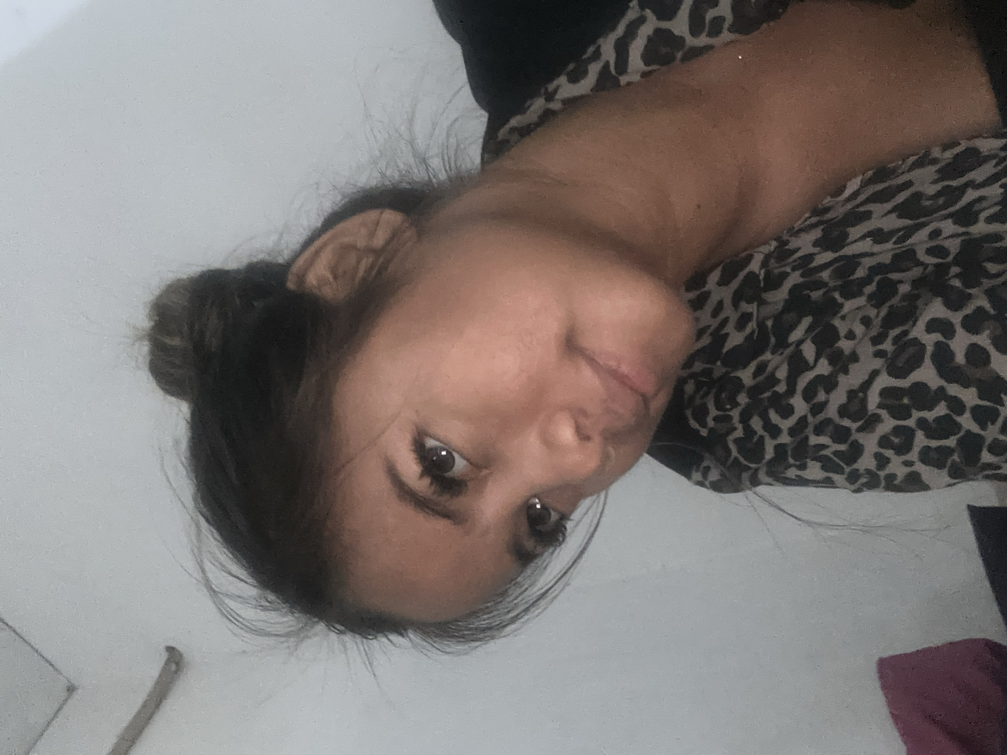 The image, rotated 90 degrees clockwise, captures an adult female indoors in North Jakarta, Indonesia. She is positioned horizontally, with her head towards the left side of the frame, looking directly at the camera with a gentle, soft smile. Her dark hair is styled in a bun, secured with a light brown scrunchie, and some wispy strands are visible around her face. She has prominent dark eyes with long, dark eyelashes.
She is wearing a top featuring a black and grey leopard print pattern. Her upper chest and neck are visible, suggesting the garment has a high neckline or is partially covered. The background consists of a plain, light-colored wall, likely white or light grey, indicating an indoor setting. In the bottom right corner of the original image, a small piece of maroon fabric is visible. Faint marks or a small hook-like object can be seen on the wall in some cropped views. The lighting appears soft, possibly from an indoor source. There is no visible text in the image.