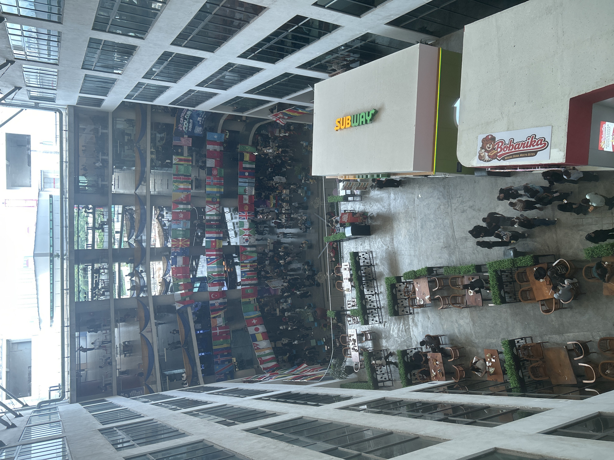 This image shows an indoor atrium-style space in Kuala Lumpur, Malaysia. There is a large gathering of people below the image, with many national flags hanging from the walls. Visible in the image are establishments like a Subway and Bobarika, along with tables and chairs set up for dining. Buildings with multiple windows are visible, suggesting an office or commercial space.