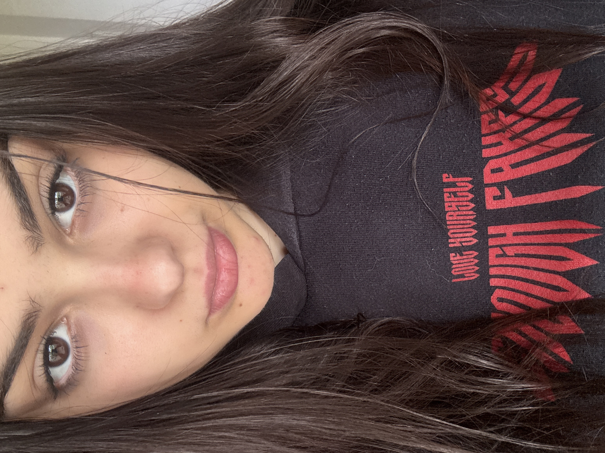 A young woman with long, dark brown hair is facing the camera. She has brown eyes and pink lips. She is wearing a dark-colored hooded sweatshirt with a red graphic design and the words "Love Yourself" printed on it. The picture is cropped close to her face and upper body. The scene does not provide any visual information about a specific location in Polena, Bulgaria or other environmental details.