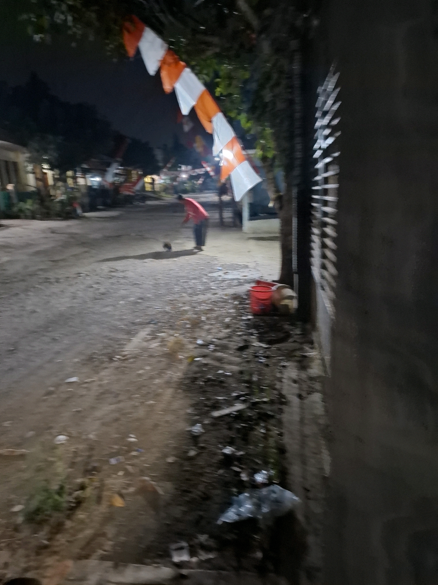 The image depicts a dimly lit outdoor scene at night in Ciheulang Tonggoh, Indonesia. In the foreground, a dirt path is littered with various debris, including plastic bags and what appear to be small pieces of trash. To the right, a textured concrete wall runs vertically, with vertical metallic bars visible in the upper portion. A dark tree with some green foliage is positioned to the left of the wall.
Further into the scene, a person wearing a red shirt and dark pants is bending over, appearing to interact with a small, dark animal, possibly a chicken or a small dog. Behind this person, Indonesian flags, alternating white and orange, are strung diagonally across the frame, indicating a festive or commemorative occasion. In the middle ground, two red buckets are placed on the ground, and another object, possibly a light-colored bucket or container, is situated next to them. Beyond this, buildings and trees are visible in the low light. The overall atmosphere is nocturnal and somewhat cluttered.