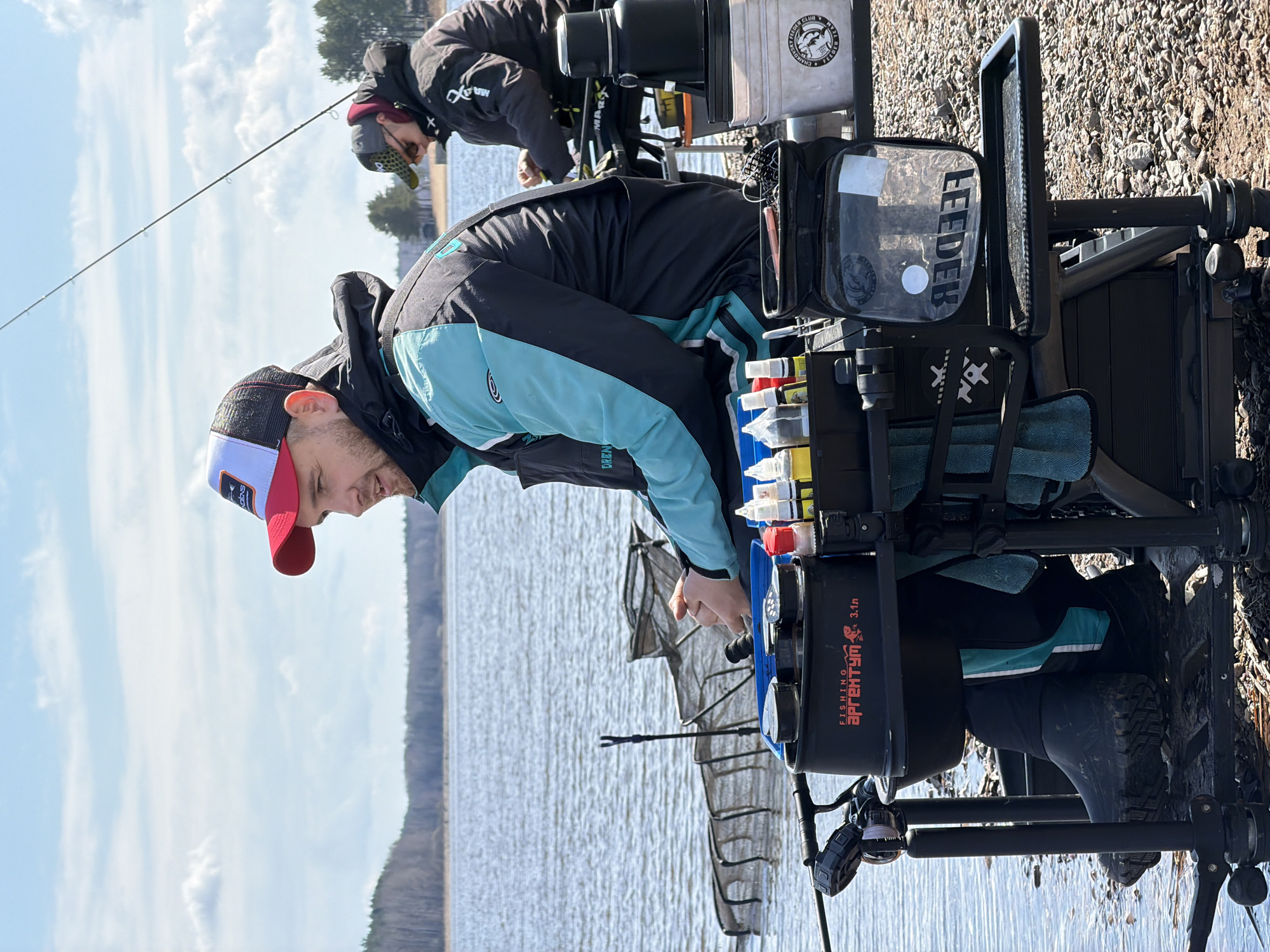 Two men are fishing on the shore of a body of water. The man in the foreground is wearing a black jacket with light blue accents and a red and white baseball cap. He is bent over a fishing station, seemingly preparing or checking something. The fishing station includes containers, compartments for tackle, and a mirror labeled "FEEDER." A second man is visible in the background, also fishing, dressed in a black jacket and hat. Fishing rods are visible extending over the water. The shore is rocky. The water extends to the horizon with a blurred background. The image was taken in the city of Chaikovsky, Russia.