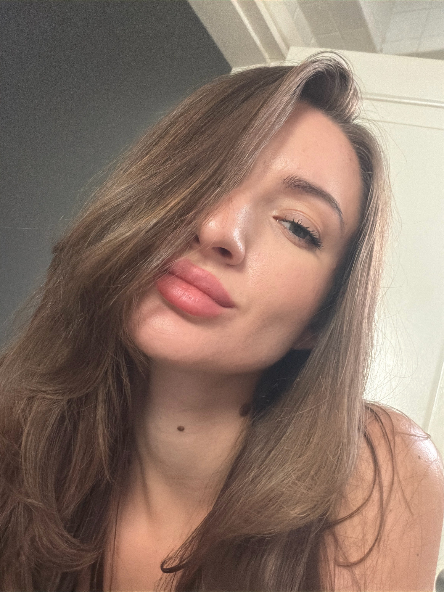 A woman with long brown hair and light skin is shown in a close-up portrait. She is looking towards the camera with her lips slightly parted. A mole is visible on her neck. The background appears to be an indoor setting with neutral-colored walls and a ceiling that suggests a doorway or partition. There is no indication of the city of Sharm El Sheikh, Egypt.