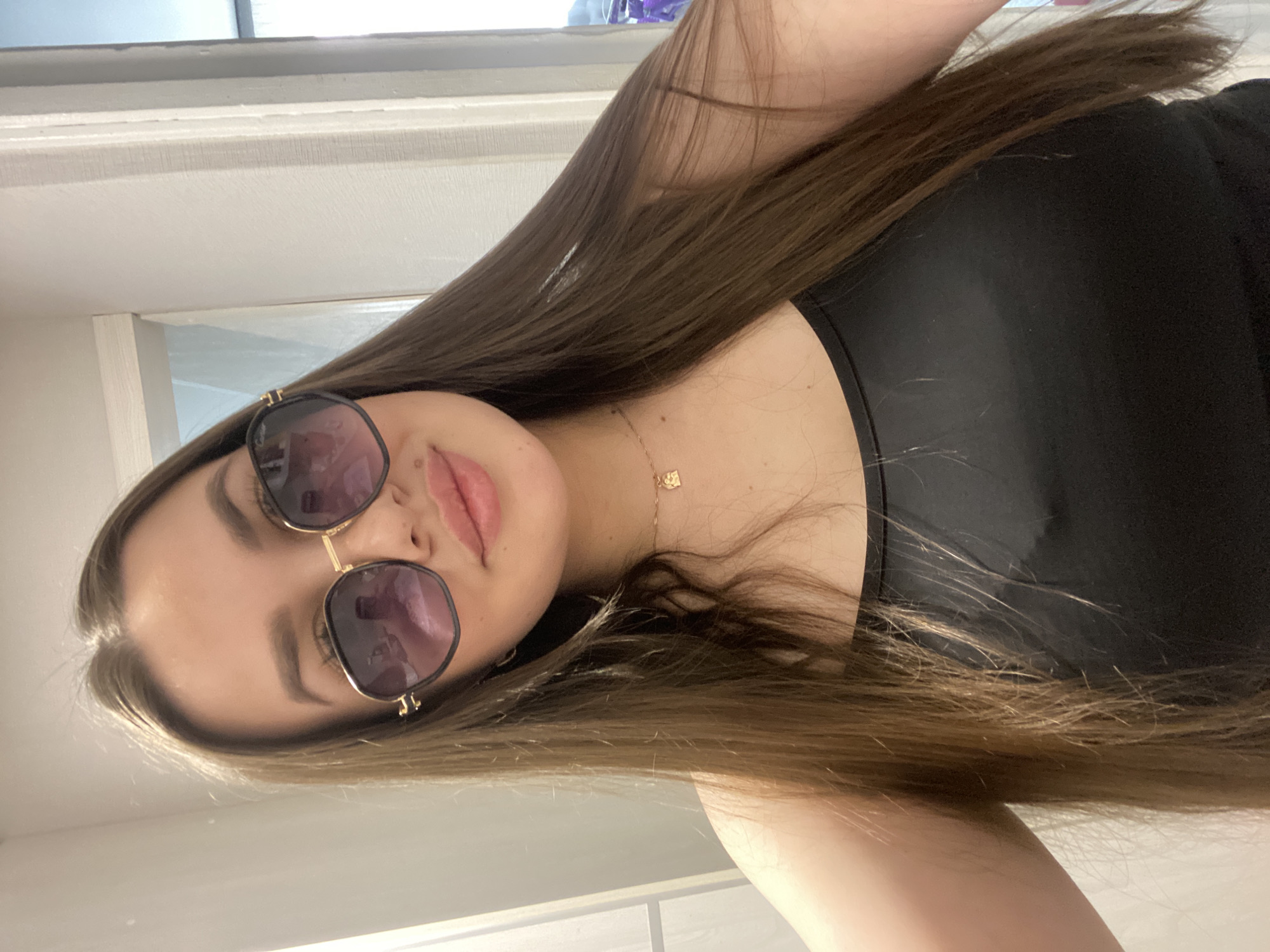 An image, rotated 90 degrees counter-clockwise, depicts a young woman in what appears to be an indoor setting in Chaikovsky, Russia. She has long, straight brown hair, parted on the side, flowing down her left shoulder and across her chest. She is wearing sunglasses with dark, possibly purple-tinted lenses and distinctive gold frames. Her lips are slightly parted, revealing a calm or subtle expression. A thin gold chain necklace with a small, square pendant adorns her neck. She is wearing a black tank top or sleeveless shirt, with her bare shoulder and upper chest visible. Her pose suggests she is taking a selfie or posing for a close-up photograph, looking towards the camera with her head tilted. The background is a light-colored, possibly white, wall or ceiling, indicating an interior space. The lighting is soft and even, highlighting her features. No visible text is present in the image.