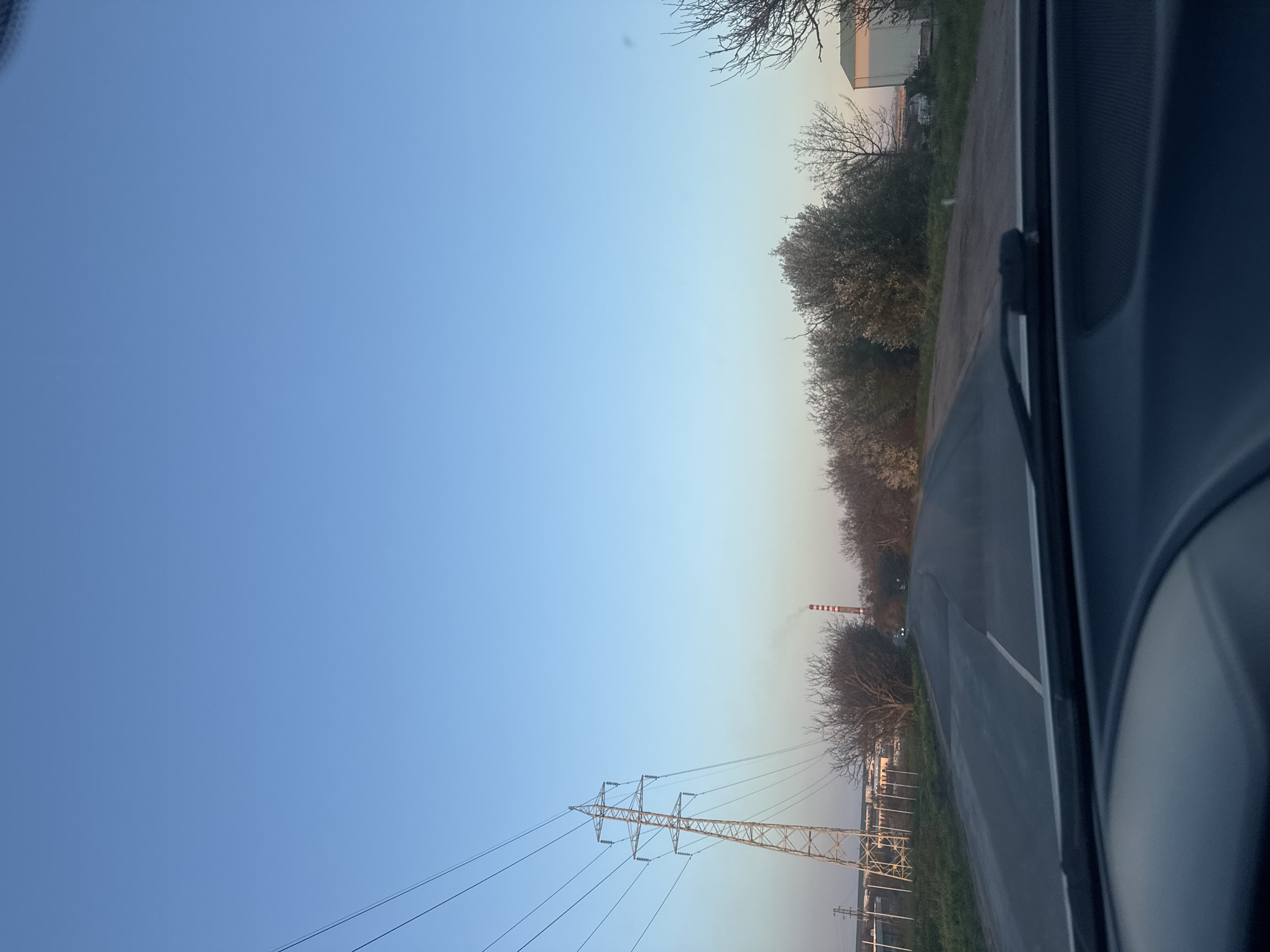 Captured from inside a vehicle, the image depicts a road scene under a clear blue sky, possibly at dusk or dawn. An asphalt road stretches into the distance, flanked by greenery and trees. A utility pole with power lines is visible in the foreground. In the distance, a red and white striped chimney or tower stands above the tree line, suggestive of industrial infrastructure. The image's location may be near Burgas, Bulgaria, as the general appearance is consistent with landscapes found in that region, though confirmation would require additional contextual information.