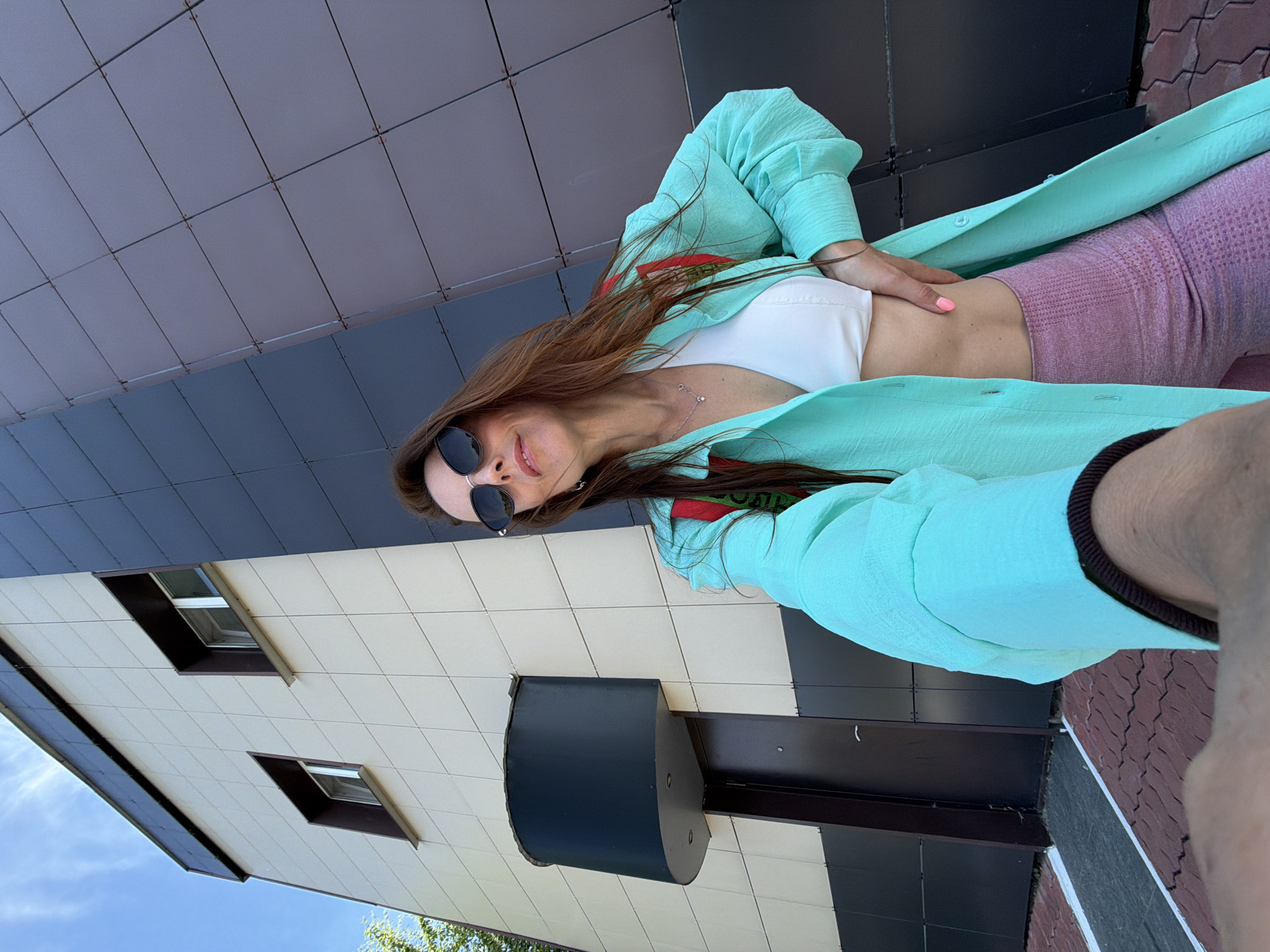 A woman with long brown hair, wearing sunglasses, a white sports bra, pink leggings, and a teal open shirt is standing outside a building. The building has a tiled facade on the left and dark panels on the right. She is holding her right hand on her hip and her left arm is extended towards the camera as if taking a selfie. The environment appears to be an urban setting with trees and a blue sky visible.