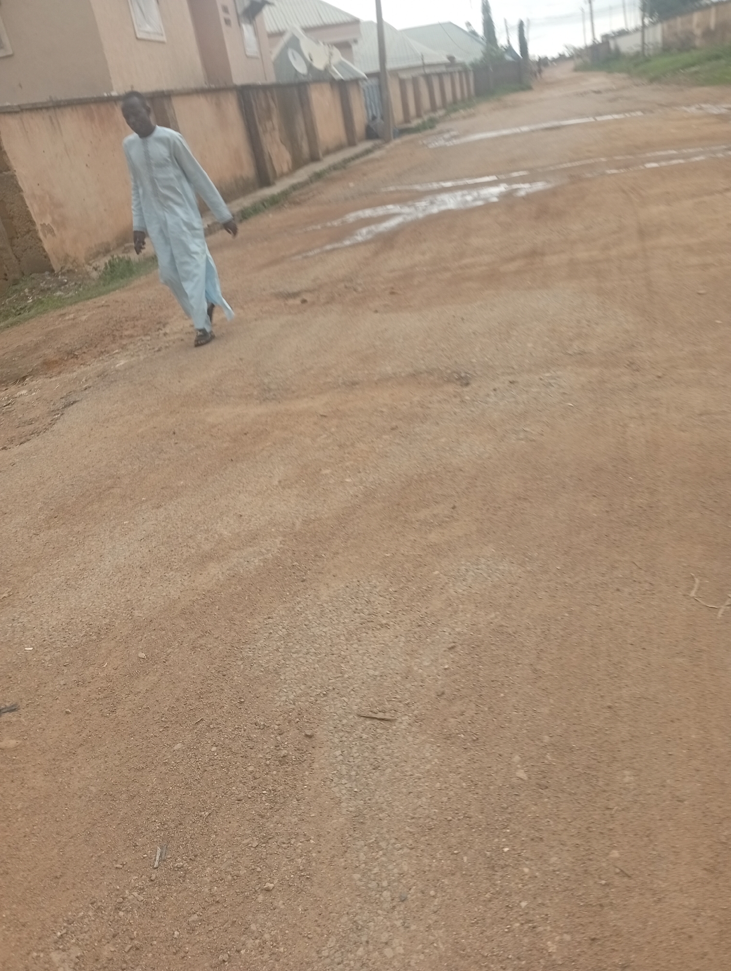 A man walks on a dirt road in Rafin Par, Nigeria. The man is wearing a pale blue traditional outfit. The road is sandy and uneven, with some puddles of water. In the background, there are buildings with beige walls and corrugated metal roofs. The scene appears to be in a residential area.
