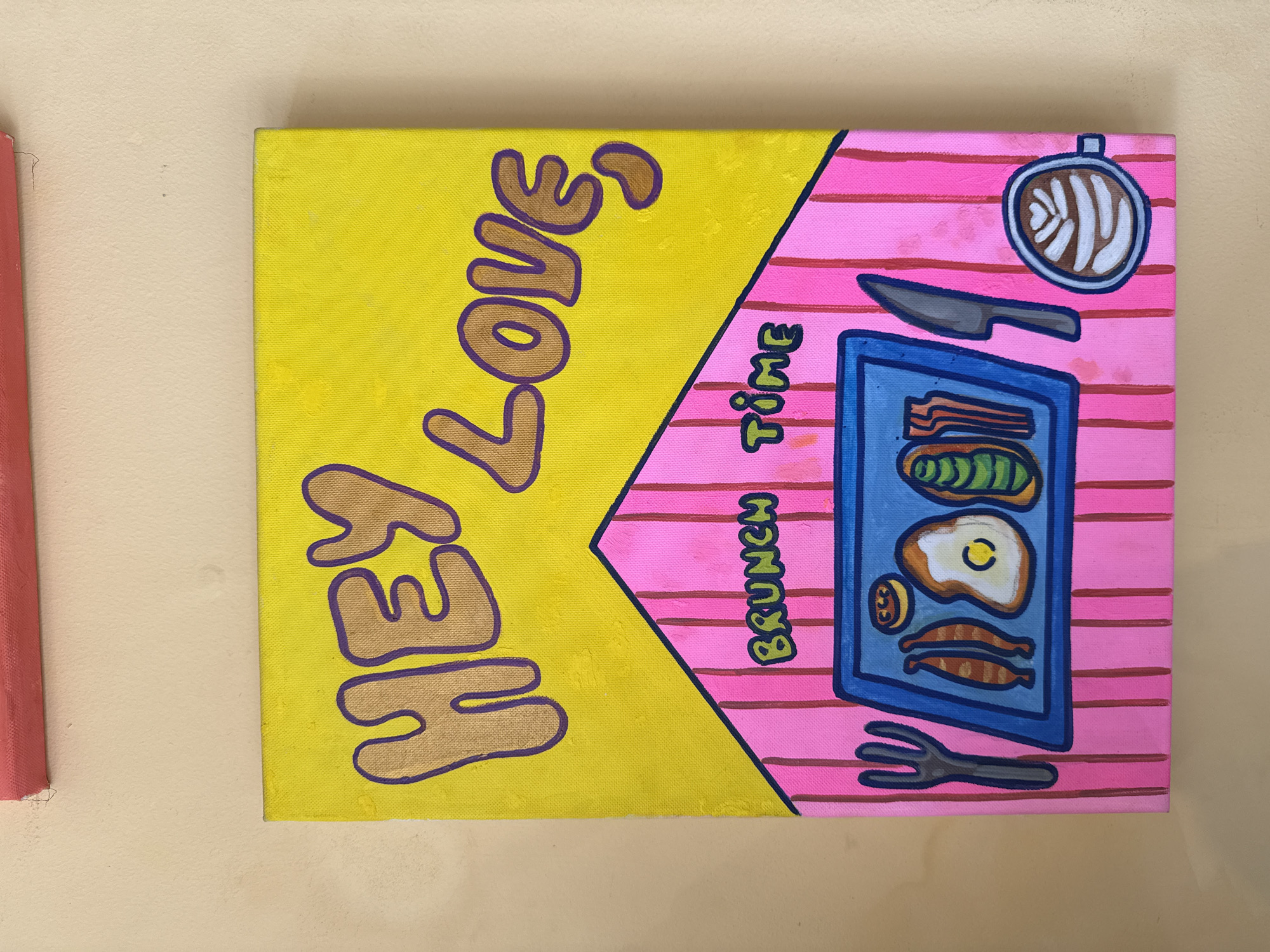 A painting on a canvas depicts the words "HEY LOVE, BRUNCH TIME" with an illustration of a breakfast platter and a cup of coffee. The text "HEY LOVE" is in gold with a purple outline and is positioned in the upper yellow section of the painting. Below this, the phrase "BRUNCH TIME" is in green with a black outline, located on a pink, striped section that resembles a roof. The breakfast platter is blue with a white outline and contains an egg, bacon, and avocado. A fork and knife are placed on either side of the platter. To the right of the platter, a coffee cup with latte art is shown. The painting is hung on a light-colored wall. There is no information provided about the location in Da Lat, Vietnam.