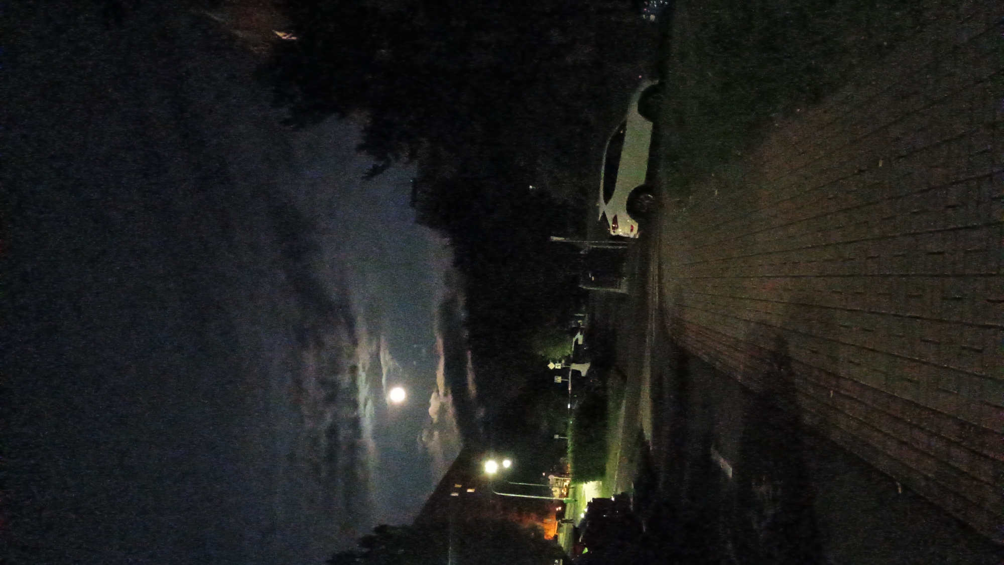 The image depicts an outdoor scene at night in Sokolov, Czechia, illuminated by the moon and artificial lights. A paved walkway made of brick or stone runs diagonally from the lower left to the upper right of the frame. To the left of the walkway, there is a grassy area and a dark-colored vehicle. Further to the left, a building with illuminated windows and a street lamp are visible.
To the right of the walkway, there is a grassy slope. A white SUV is parked on this slope, angled towards the right. Beyond the SUV and the walkway, dense trees and foliage create a dark, shadowy background. The sky above is dark with patches of clouds, and a bright, full moon is visible high in the sky, partially obscured by clouds. The overall impression is a quiet, nocturnal urban or suburban landscape.