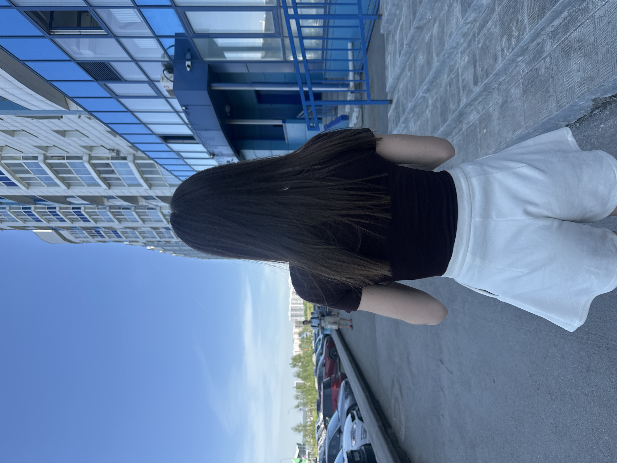 The image, originally rotated ninety degrees clockwise, captures an outdoor urban scene on a clear, sunny day. The primary subject is a person viewed from behind, walking away from the camera. This individual has long, dark brown hair and is wearing a black short-sleeved top and white shorts or a short skirt.
To the left of the person (when the image is correctly oriented), a large, modern building occupies the background. Its facade features numerous square and rectangular windows, many reflecting the bright blue sky, interspersed with sections of blue paneling. A distinct blue metal structure, possibly a railing or exterior staircase, is visible against one of the building's blue wall sections.
To the right of the person, a paved ground surface, resembling a sidewalk or parking lot, extends into the distance. Further beyond this, a row of parked cars of various colors lines a street or lot. In the far background, tiny figures of other people are visible, along with distant buildings and green trees under a clear blue sky. The overall scene suggests a vibrant urban or suburban setting during daylight hours. No visible text is present in the image.