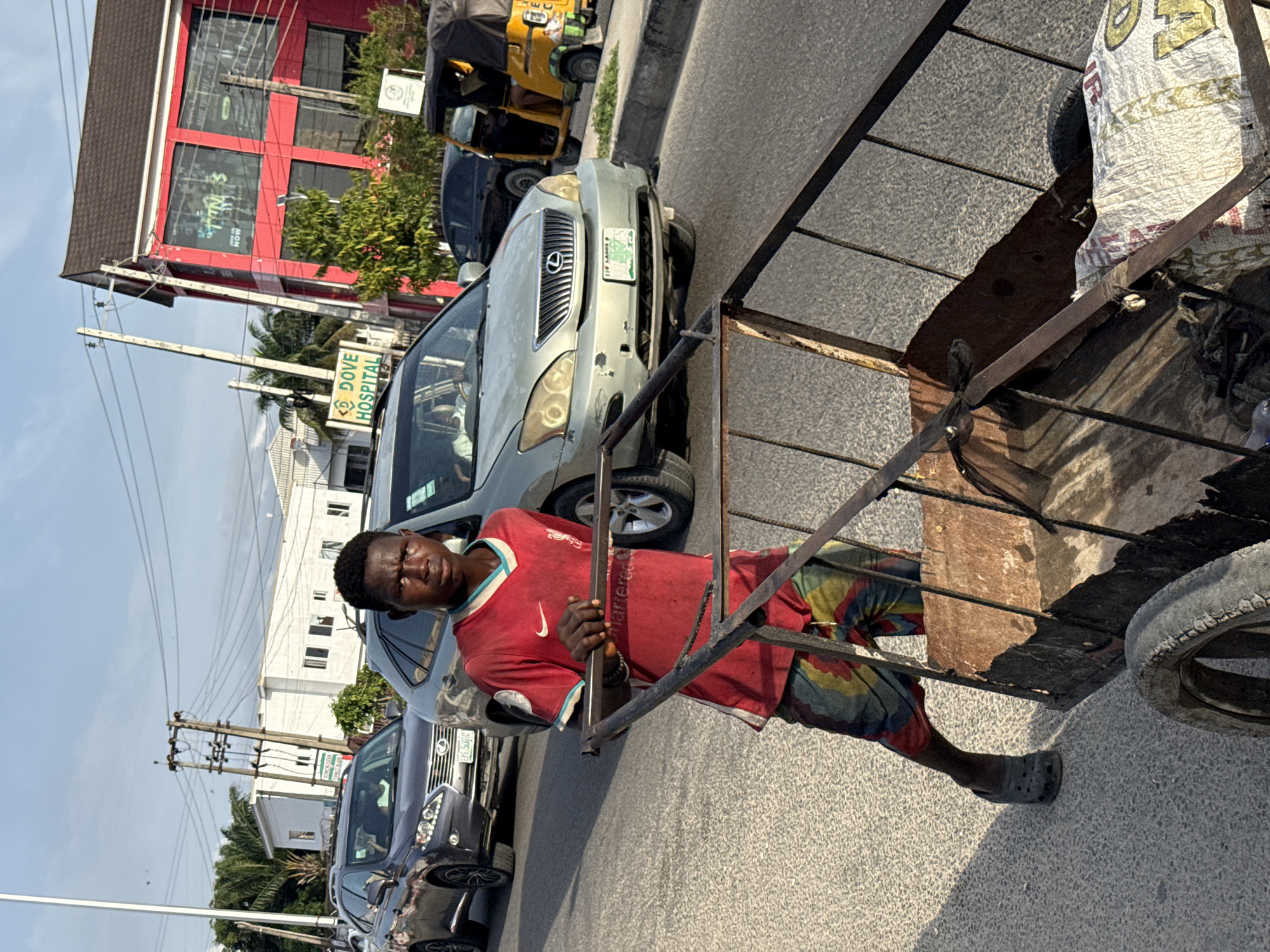 A man in a red shirt and multicolored shorts pulls a cart on a street in Ado, Nigeria. He wears a grey Croc on his right foot. The cart has a metal frame and wheels, with a wooden and cloth-covered load. Cars are parked along the street, including a silver Lexus with license plate "116 LAGOS F". A yellow tricycle is visible further up the street. Buildings are visible on the left, including a red building and a building with a "DOVE HOSPITAL" sign. Overhead power lines run along the street. The sky is light blue.
